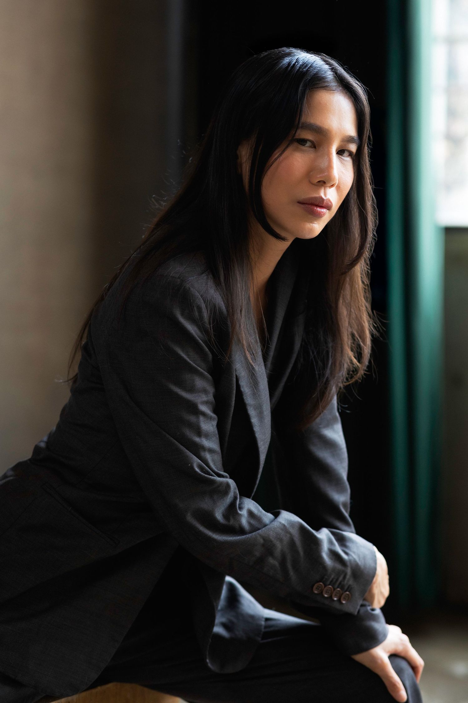 Woman in black blazer sits contemplatively in moody lighting.