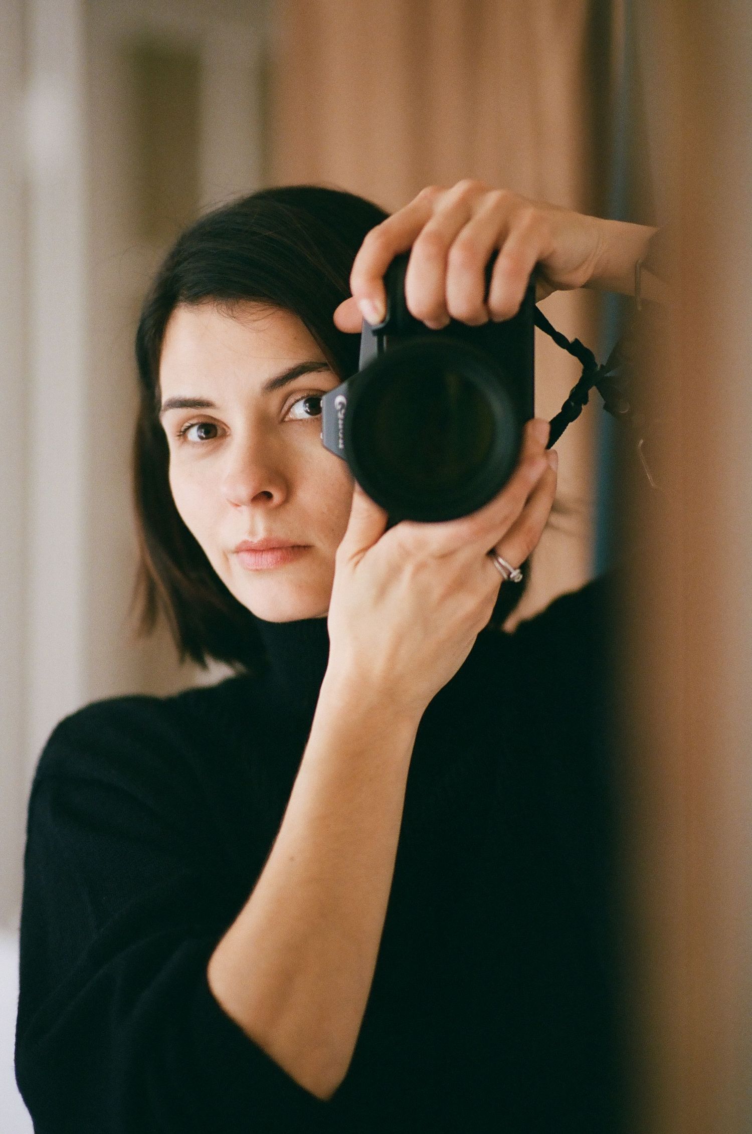 A young woman with short dark hair holds a DSLR camera up to her eye, wearing a black turtleneck sweater.