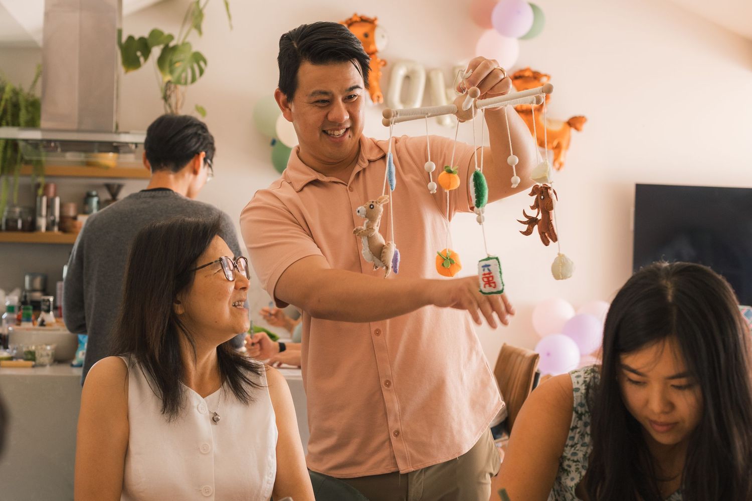 A smiling man in a pink shirt holds decorations at a festive indoor gathering with friends nearby.