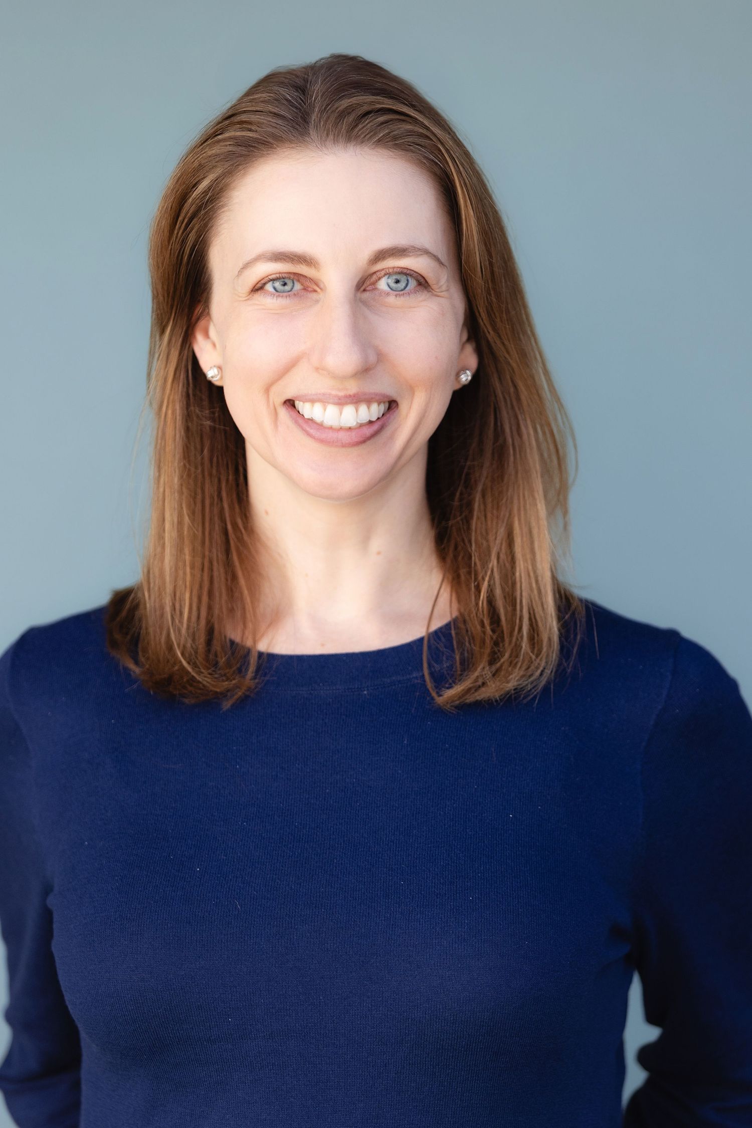 Smiling woman with brown hair and blue eyes wearing a navy blue sweater against a light gray background.