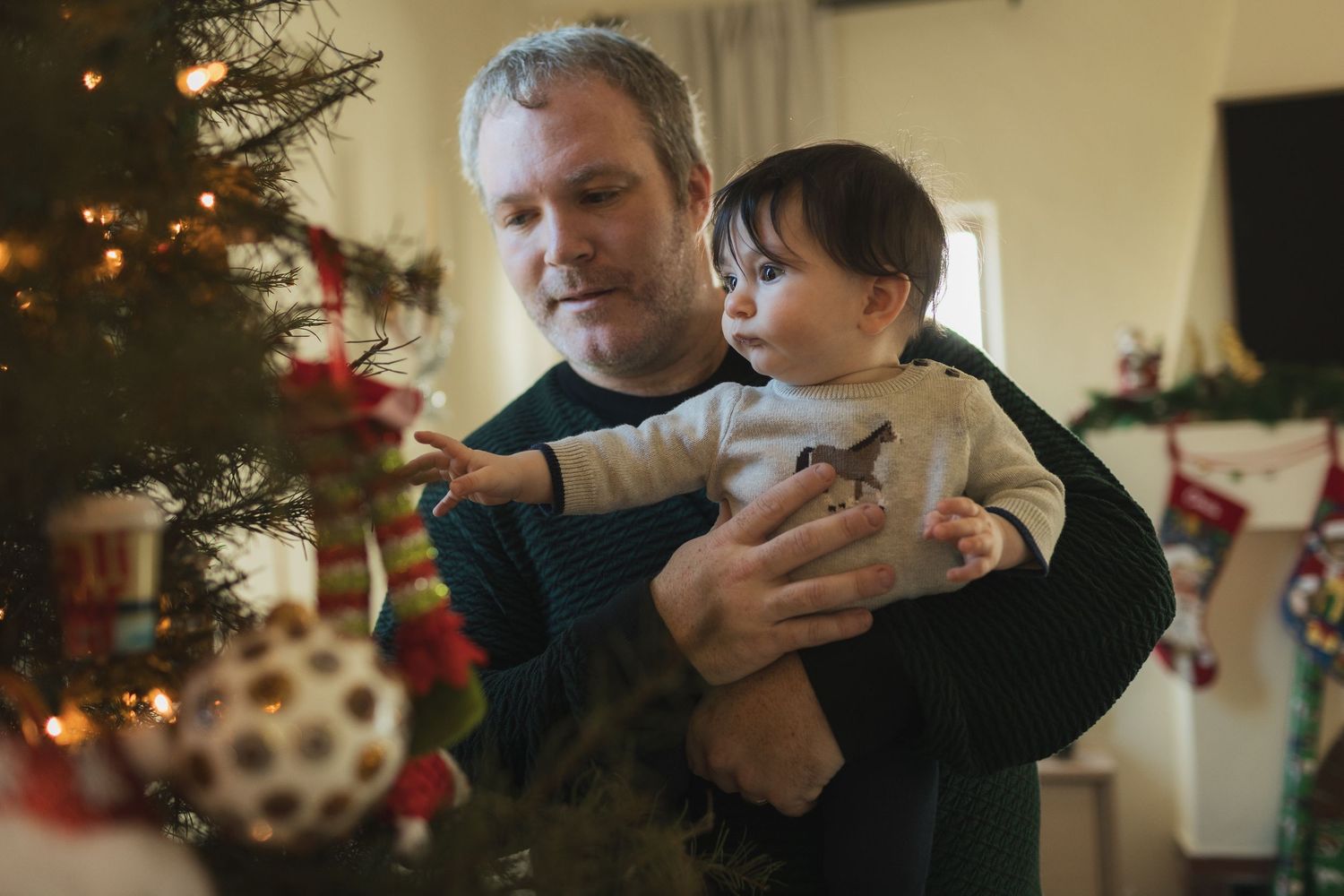 Father holding baby while decorating Christmas tree with ornaments, stockings hanging in background.