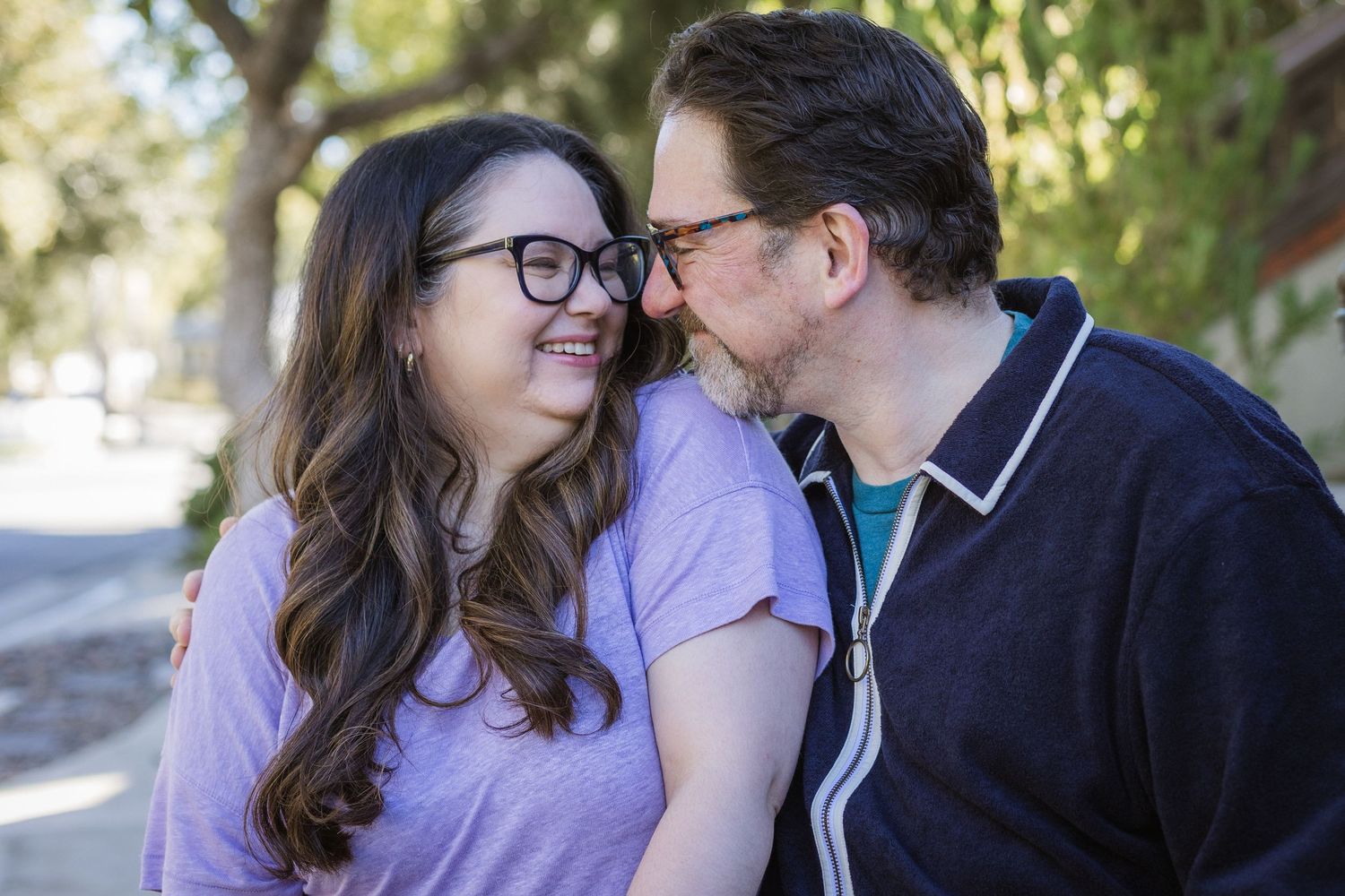 A smiling couple shares a tender moment outdoors, man kissing womans cheek, woman wearing glasses and lavender shirt.