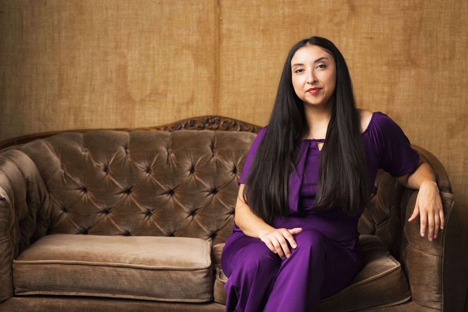 Woman in purple dress sitting elegantly on a vintage brown tufted sofa against a golden textured wall.