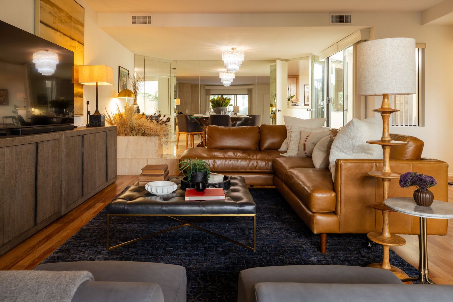 Modern living room with tan leather sectional sofa, dark wood coffee table, navy rug, and warm ambient lighting.