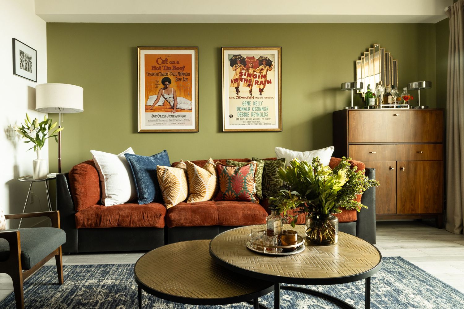 Olive green living room with rust velvet sofa, colorful pillows, vintage posters, and round coffee table with flowers.