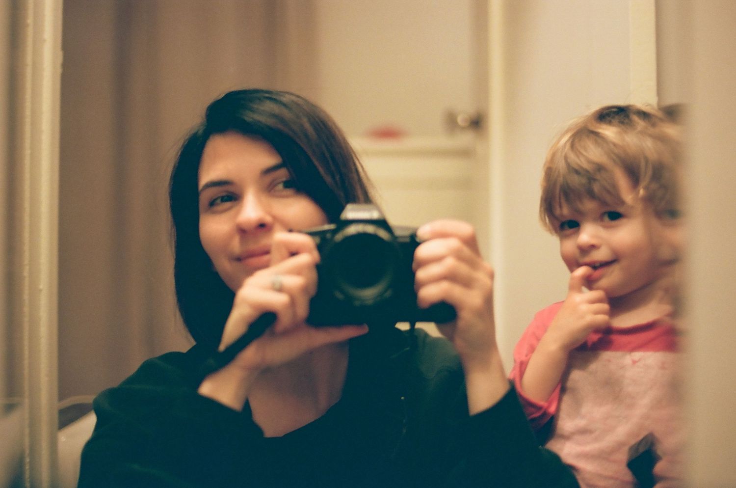 Woman taking mirror selfie with camera while young toddler girl smiles beside her in warm indoor setting.