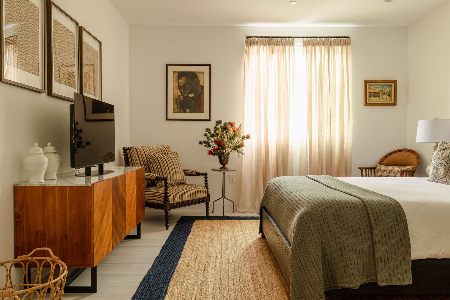 Elegant hotel bedroom with wood dresser, TV, striped armchair, neutral tones, framed art, and natural light through sheer curtains.