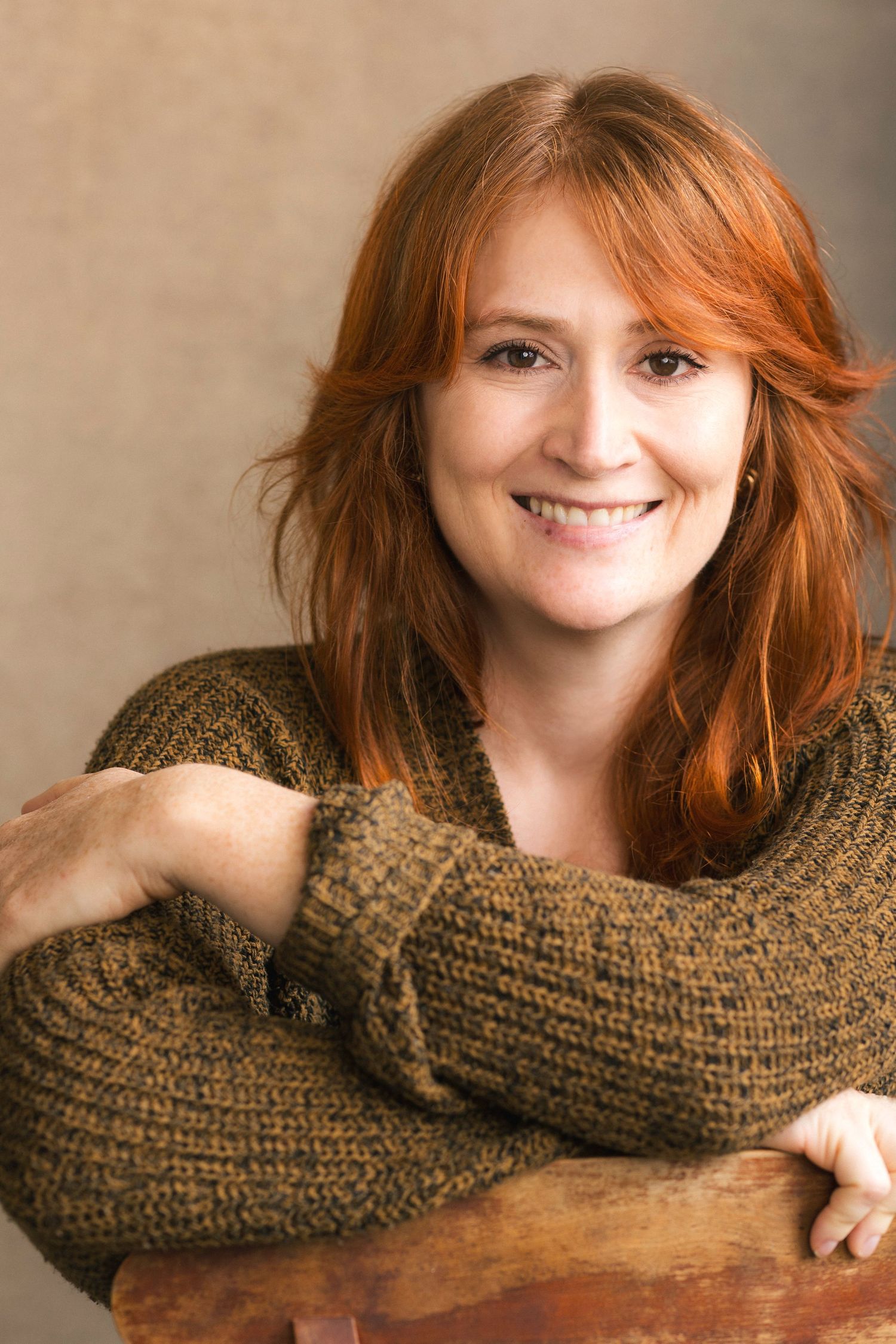 Smiling redhead woman in brown knit sweater posing with arms crossed on wooden surface against neutral background.