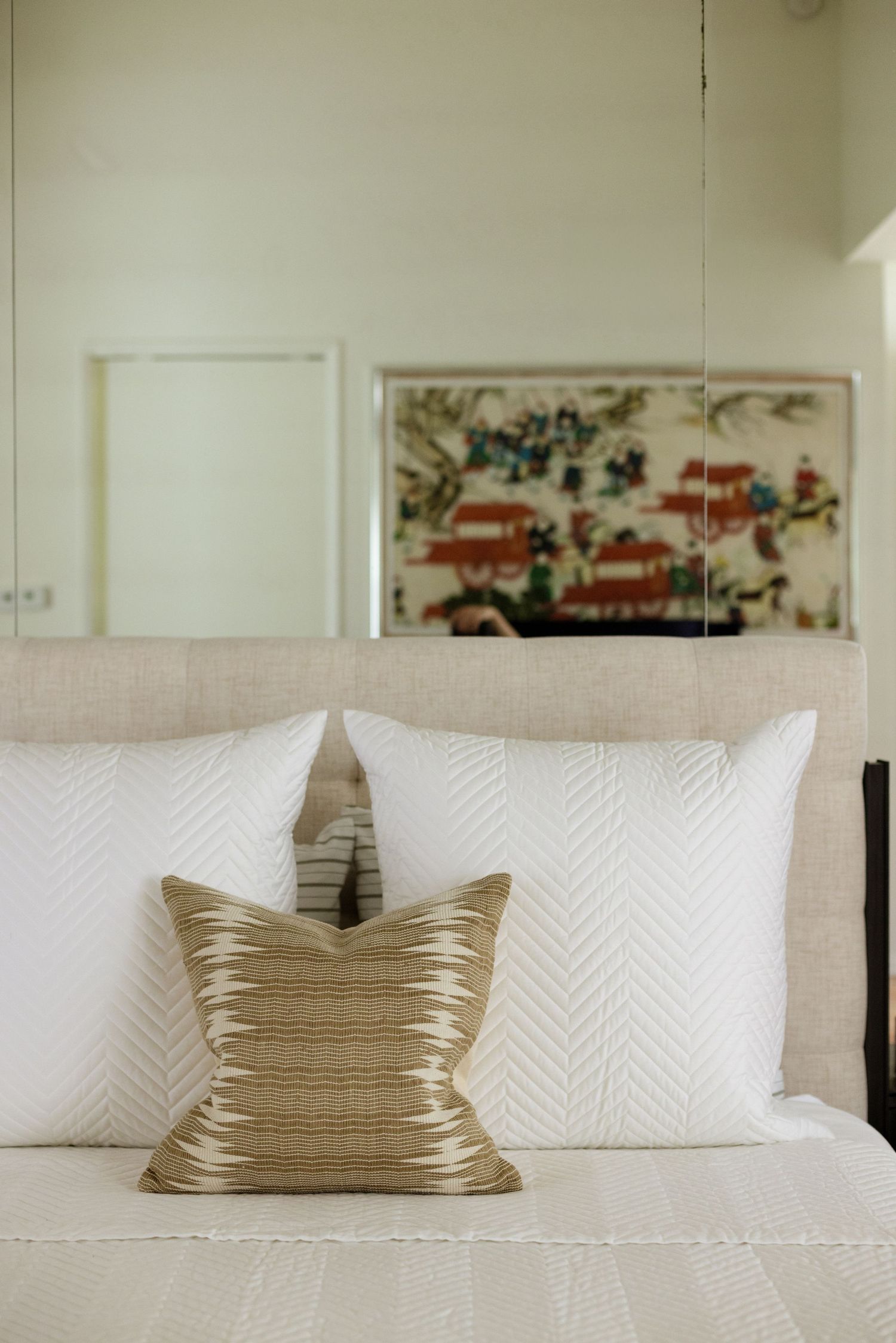 Elegant bedroom with white textured pillows and a gold accent cushion, featuring Asian artwork above a neutral upholstered headboard.