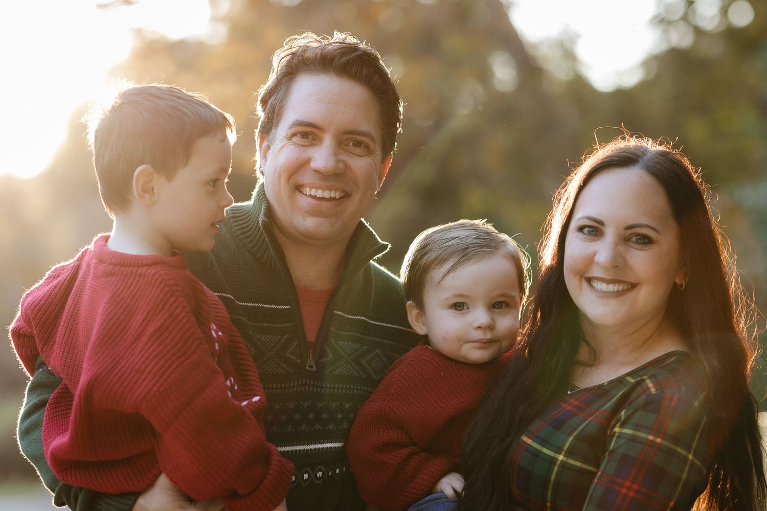 Happy family of four posing outdoors in autumn light, parents holding two young children wearing red holiday outfits.