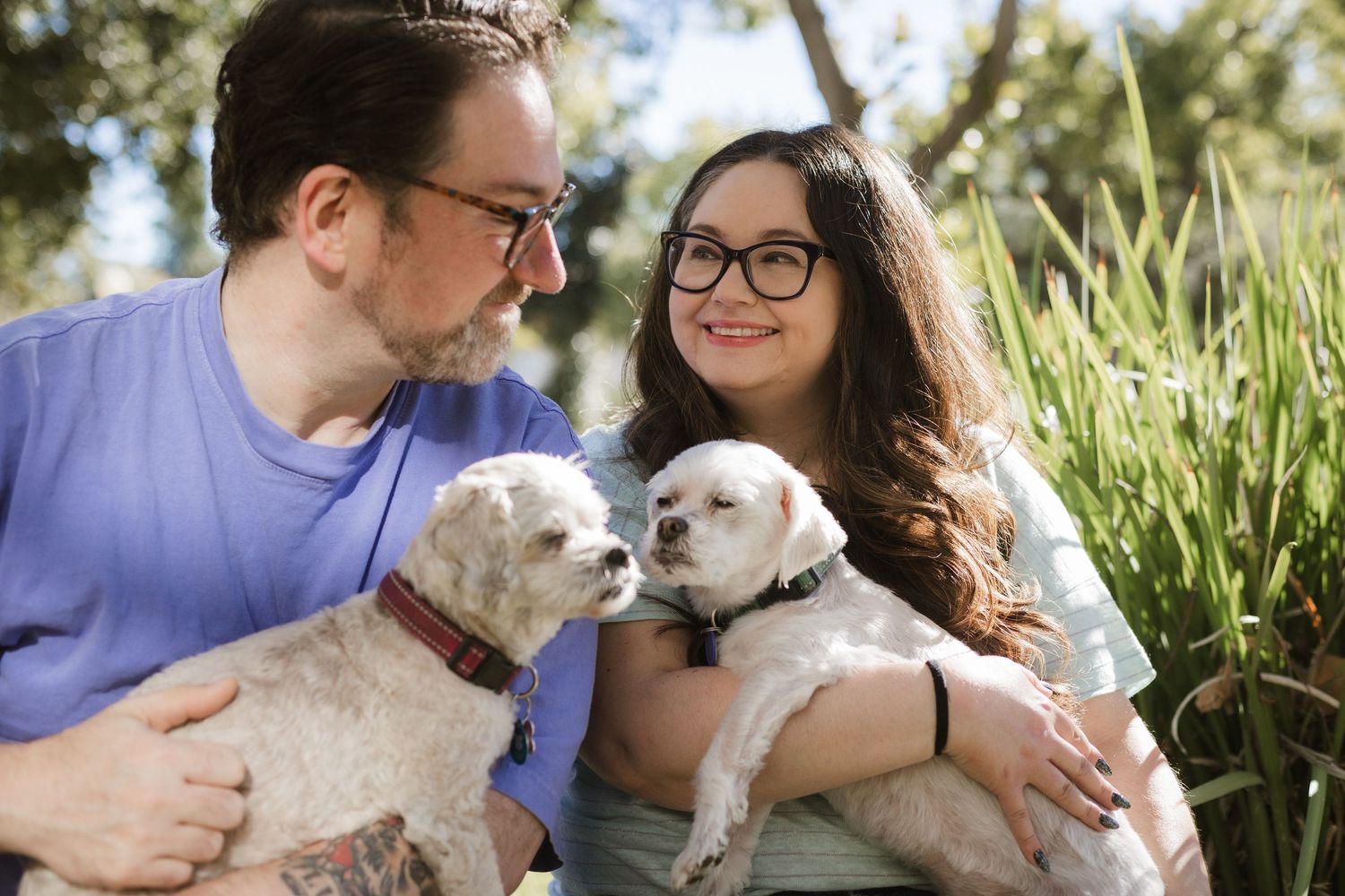Couple cuddles with their small white dogs outdoors in garden setting.