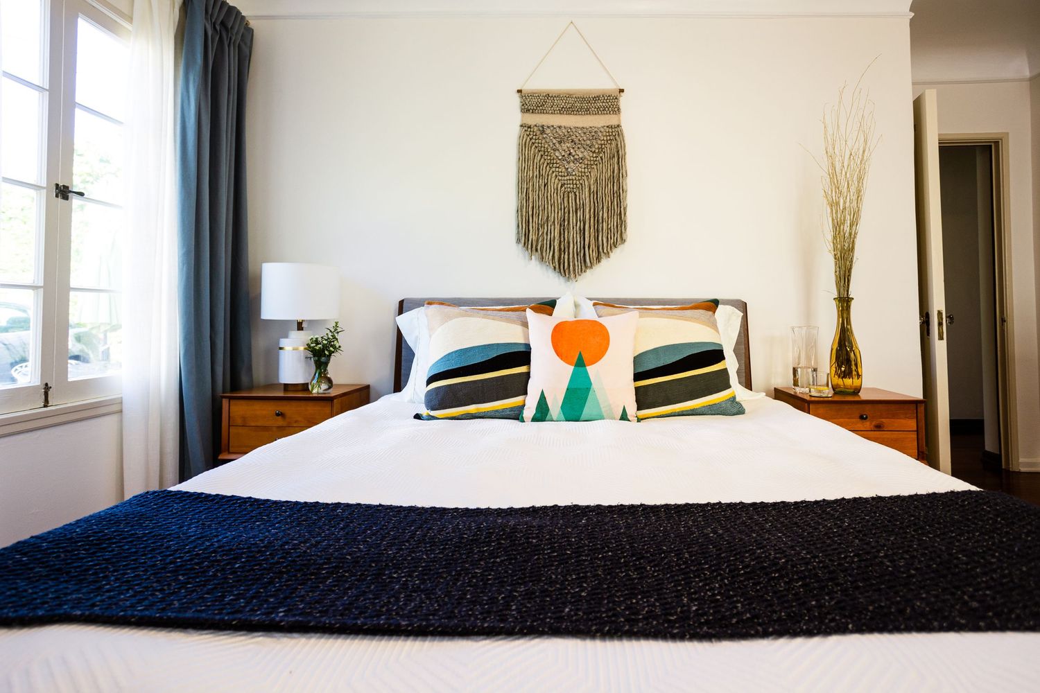 Boho bedroom with navy throw, colorful pillows, macrame wall hanging, and wooden nightstands.