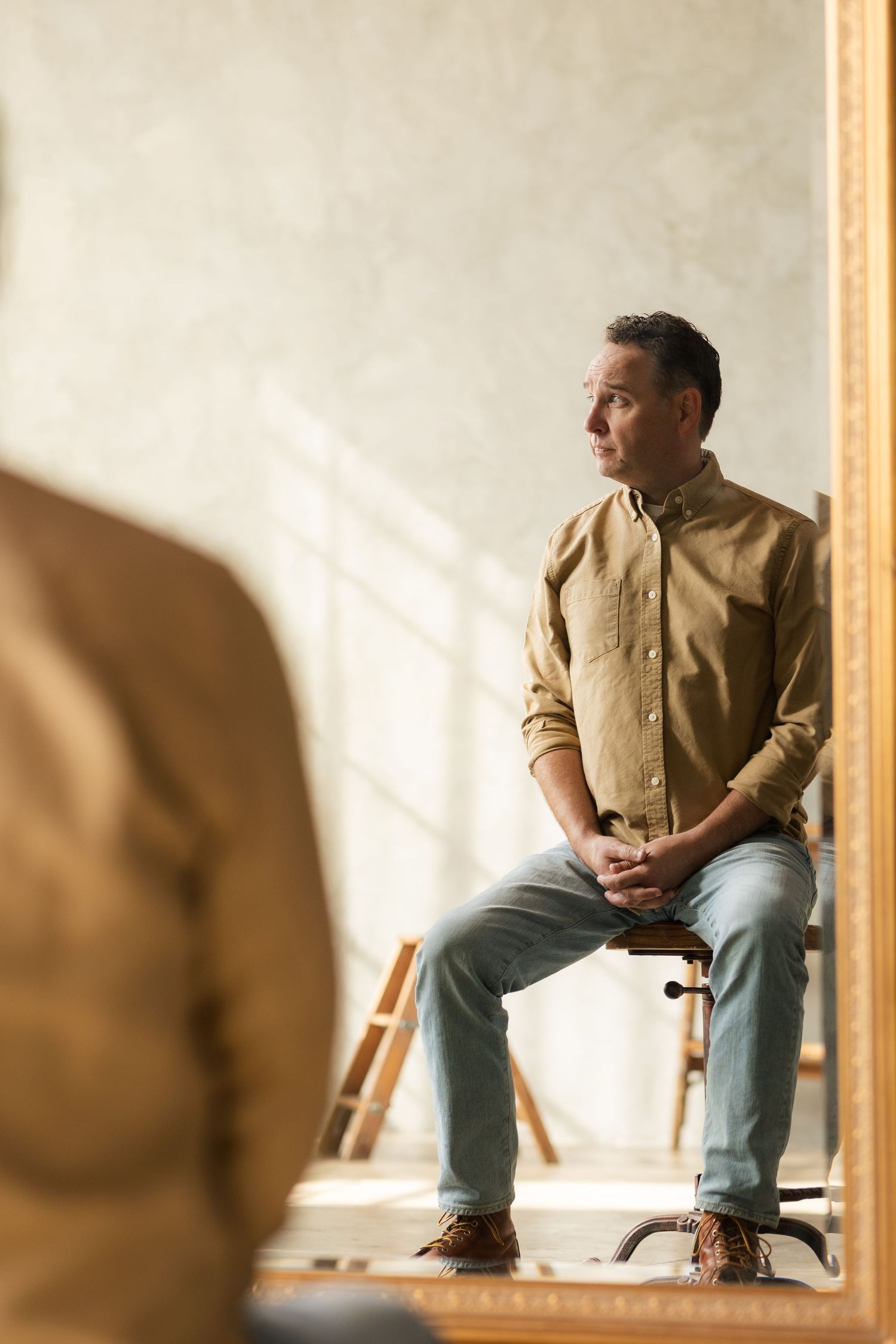 Person in tan jacket sitting on wooden chair with artistic mirror reflection.