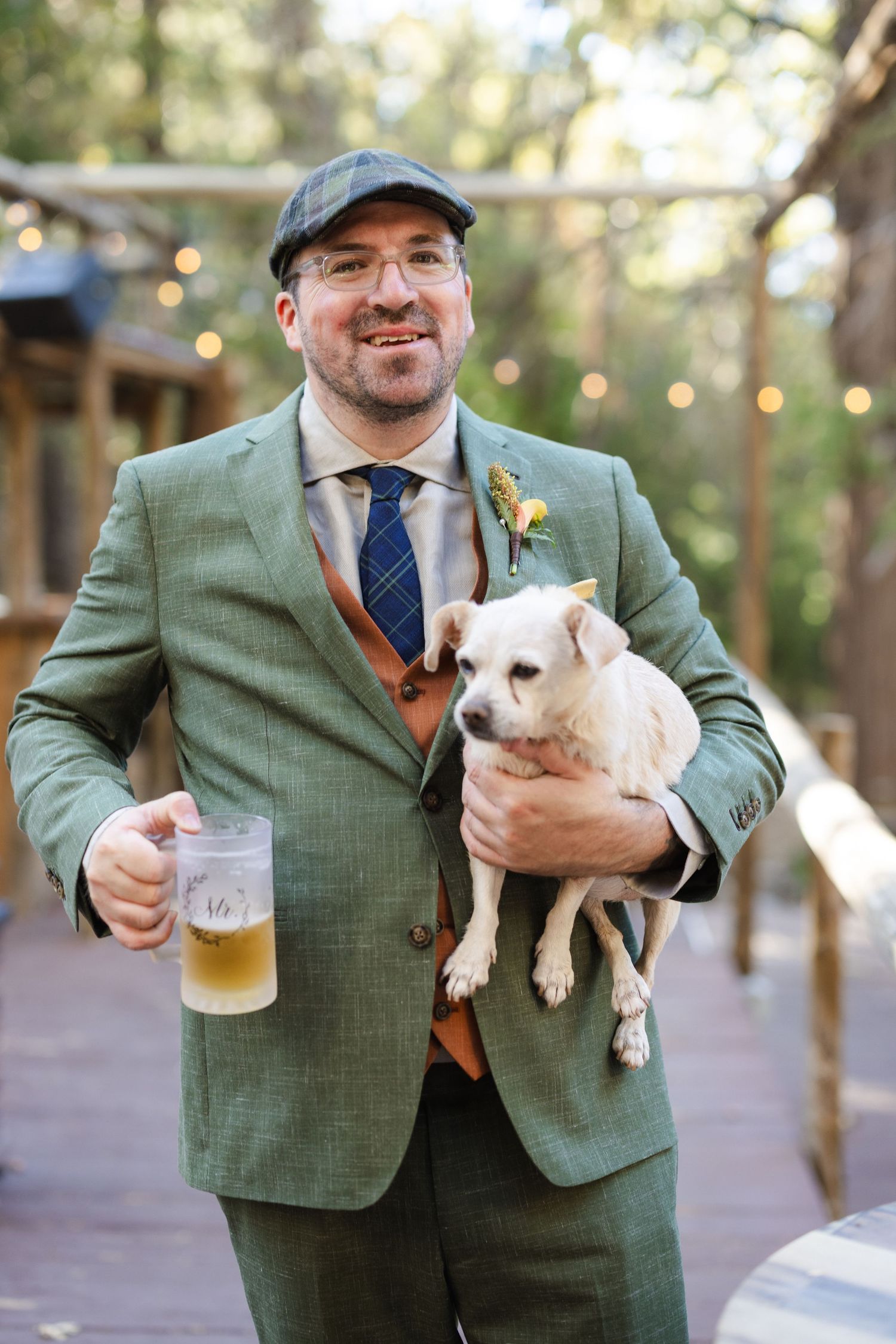 Person in green suit jacket holding small white dog while enjoying a beer outdoors.