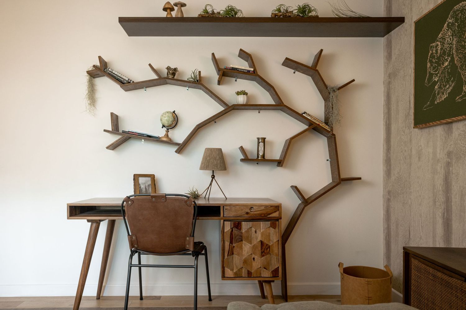 A creative tree-shaped wooden bookshelf mounted on a wall above a mid-century modern desk with a leather chair.