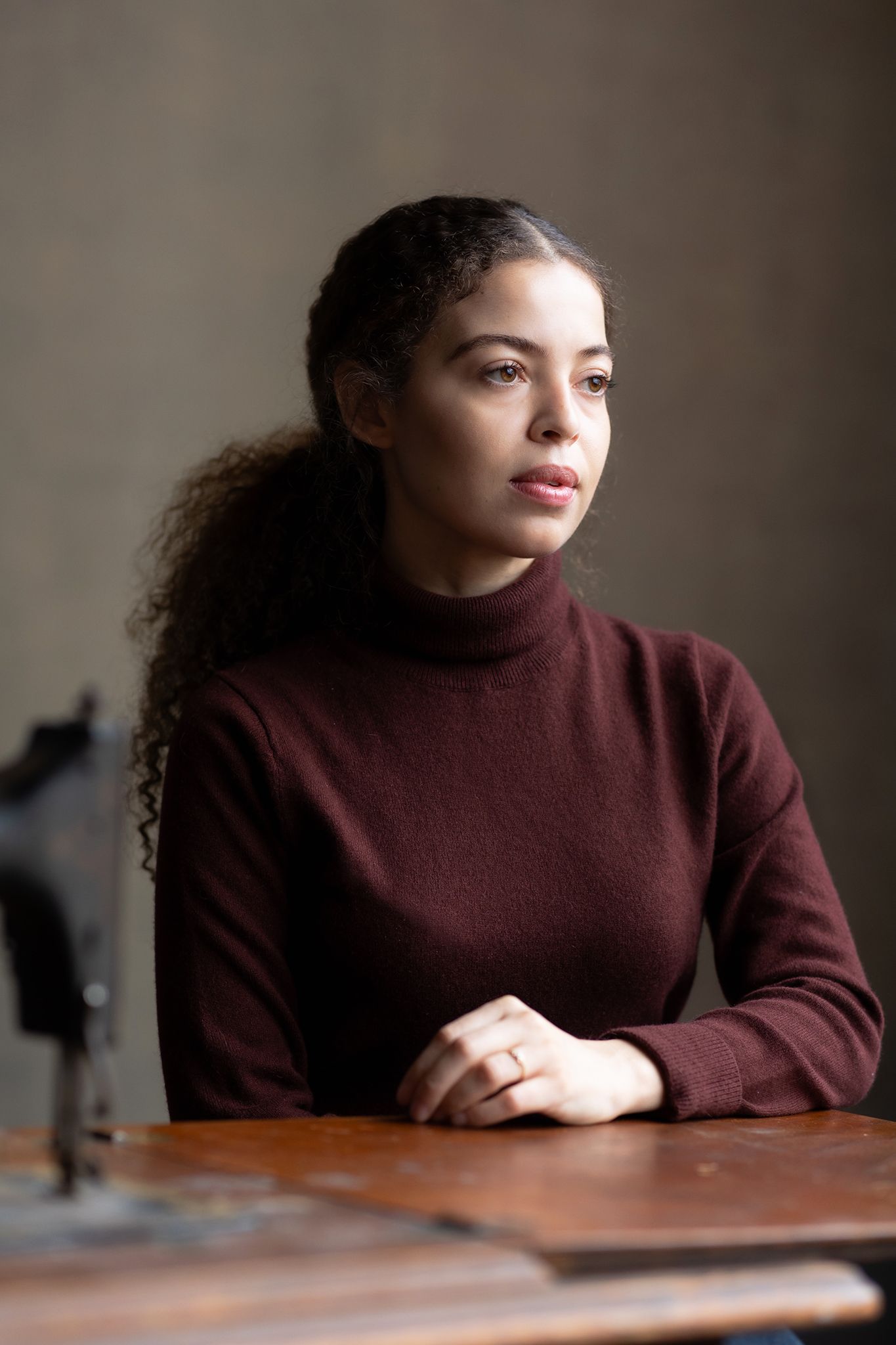 Person in burgundy turtleneck sweater sits at wooden table in moody lighting.