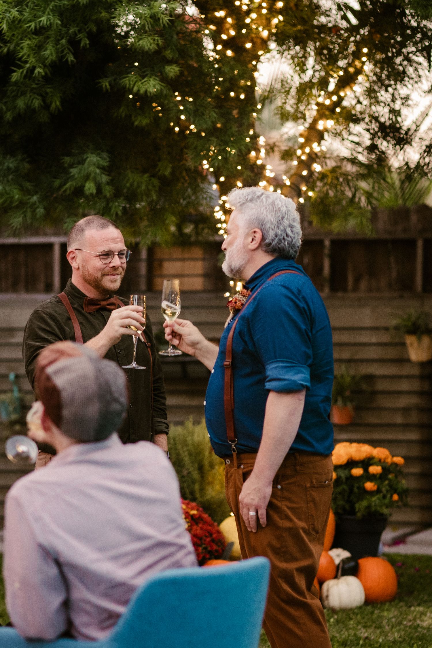 People toasting with champagne glasses at a backyard party with string lights and pumpkins.
