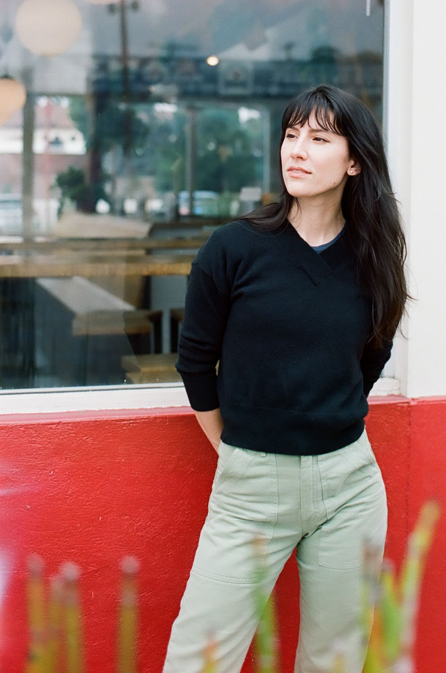 Young woman with dark hair in black sweater and light pants leaning against a red wall outdoors.