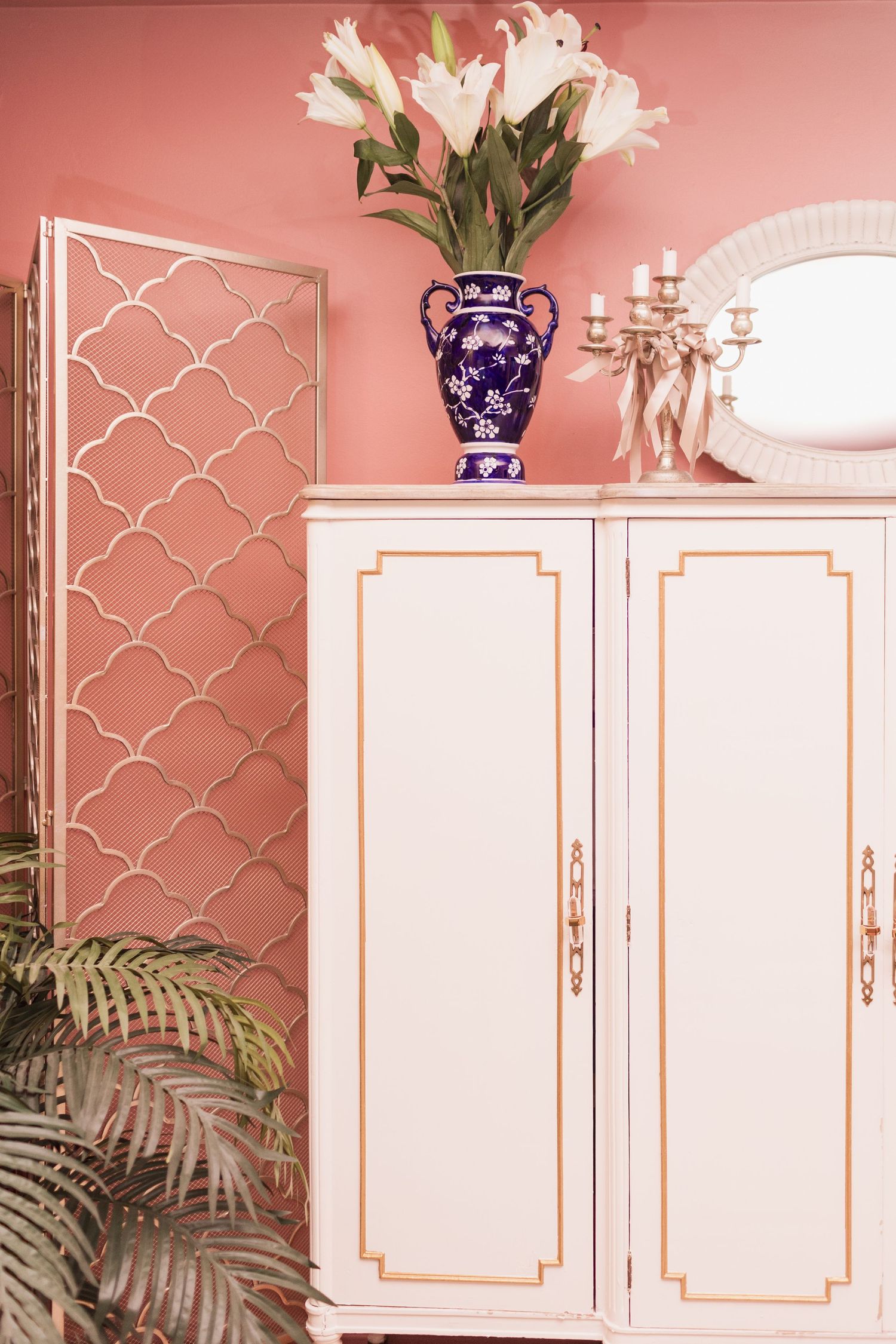 White vintage armoire with gold trim, blue floral vase, candelabra, and mirror on pink wall with tropical plants.