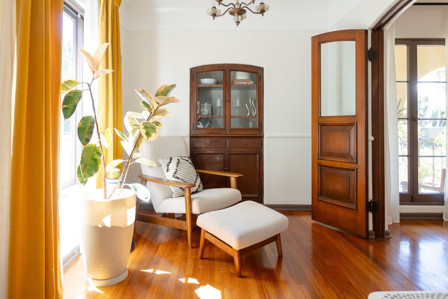 Bright living room with hardwood floors, yellow curtains, wooden chair with ottoman, antique cabinet, and tropical plant.