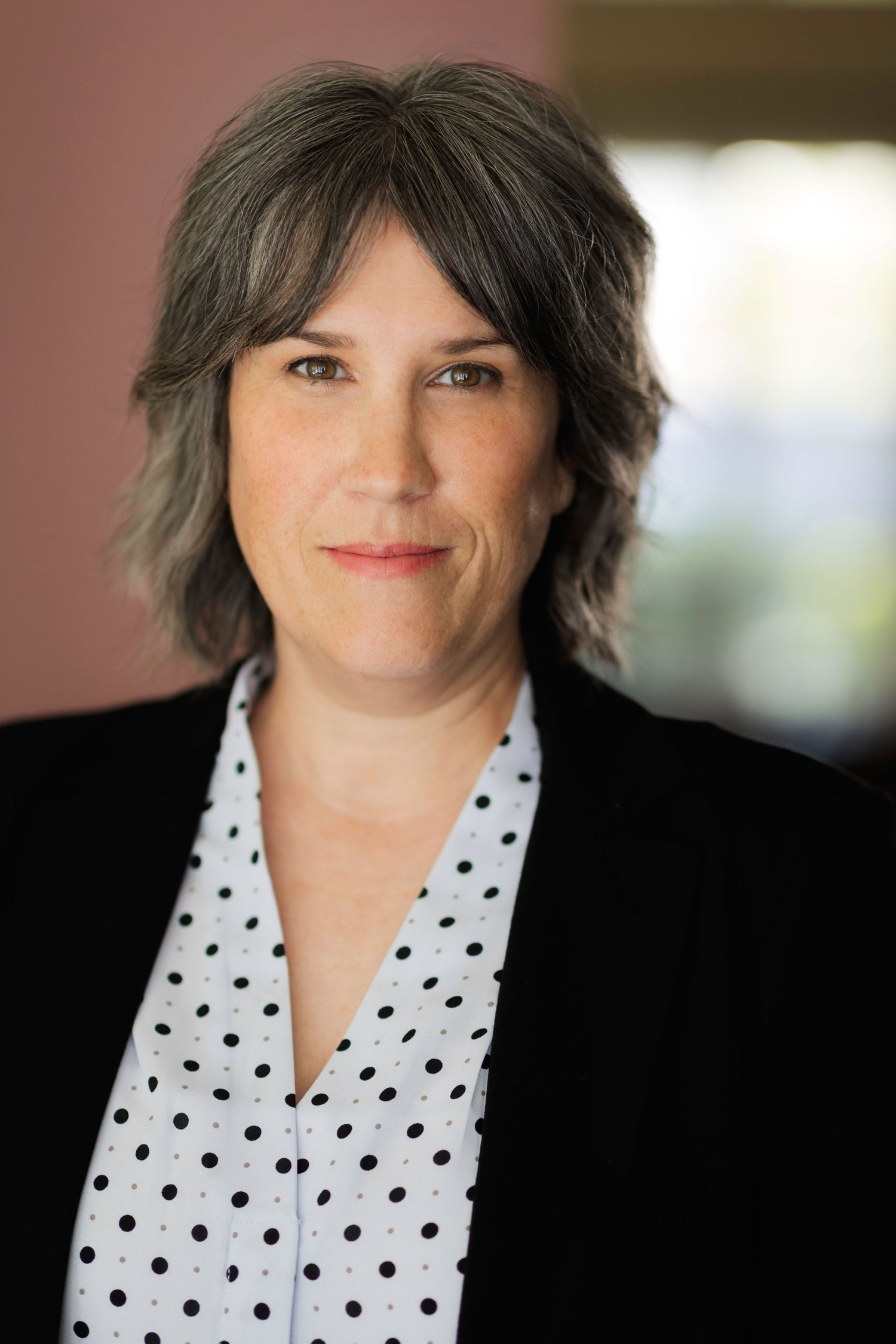 Professional headshot of a woman with gray hair wearing a black blazer and white polka dot blouse, smiling confidently.