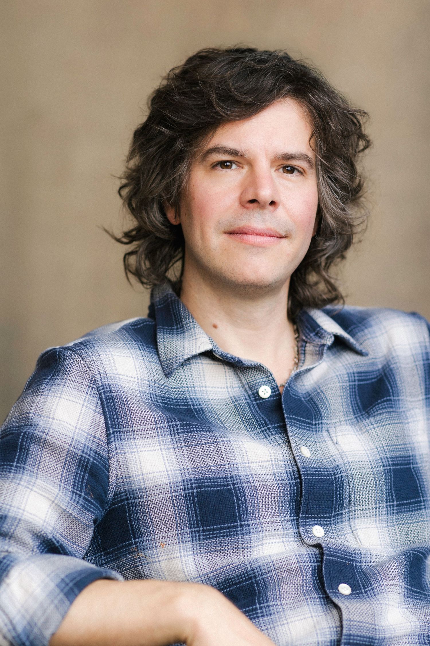 A man with curly dark hair wearing a blue plaid flannel shirt, smiling softly against a neutral background.
