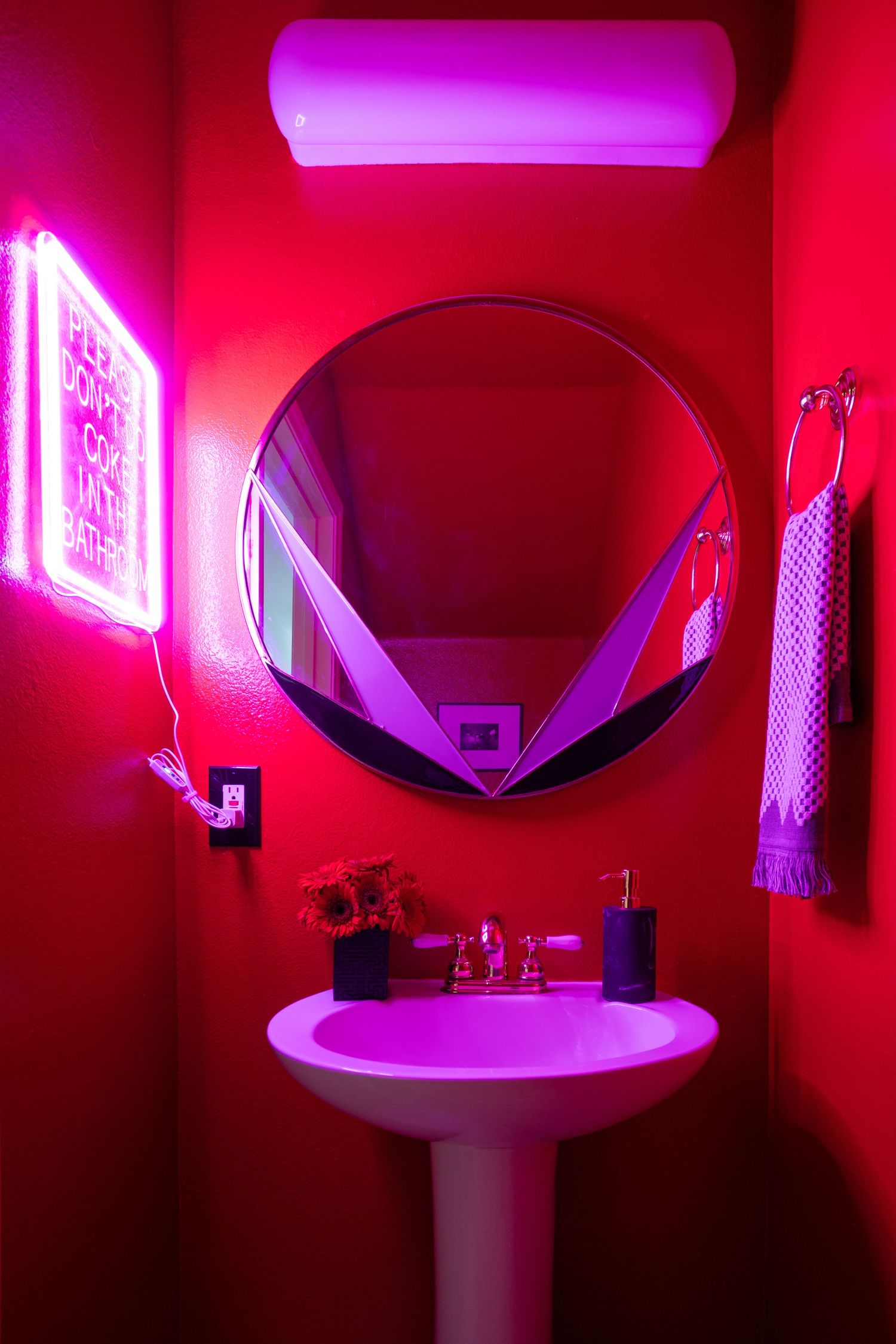 Vibrant red bathroom with neon pink lighting, round mirror, pedestal sink, and glowing neon sign wall art.