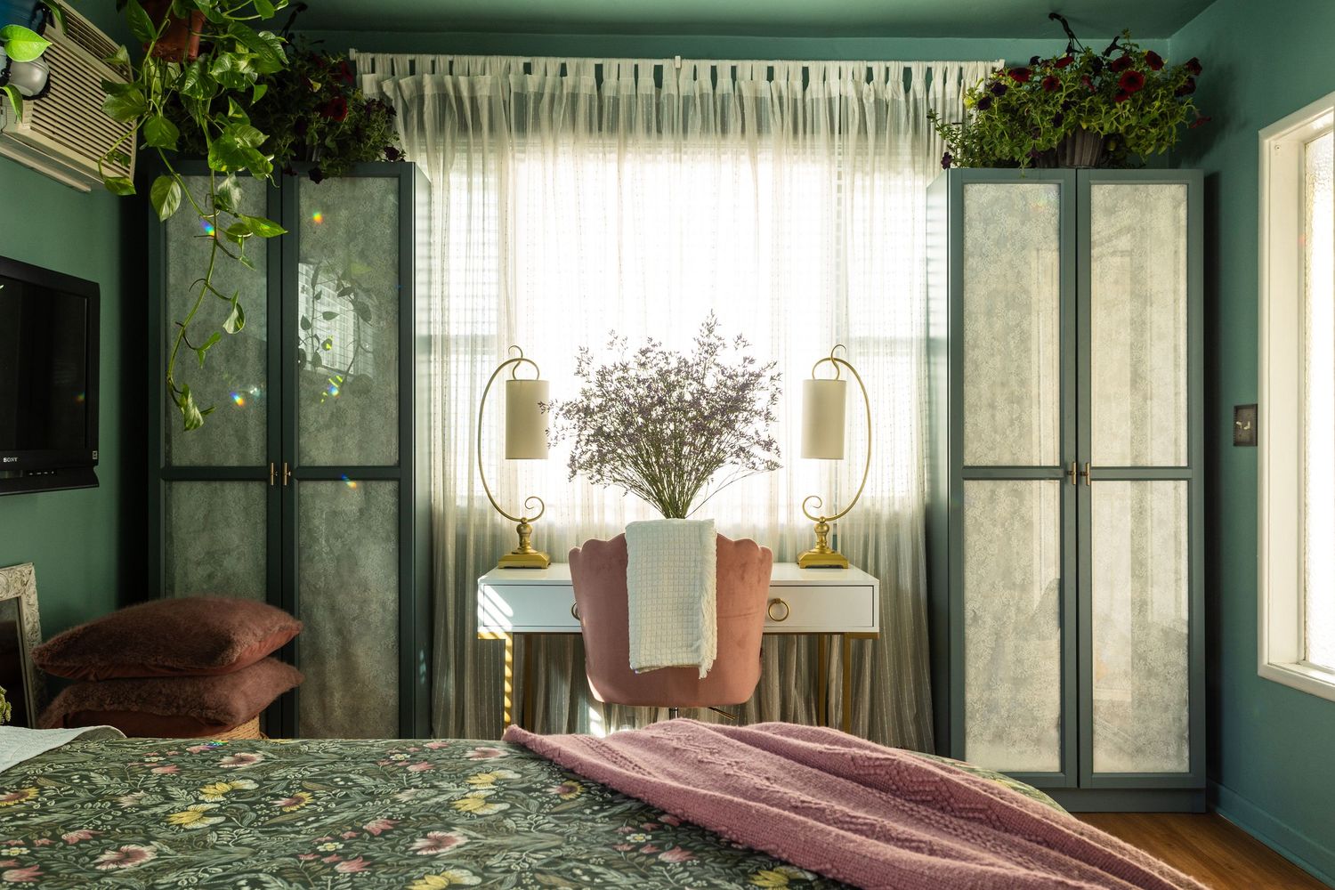 Cozy bedroom with green walls, mirrored wardrobes, floral bedding, potted plants, and a white vanity with gold lamps.