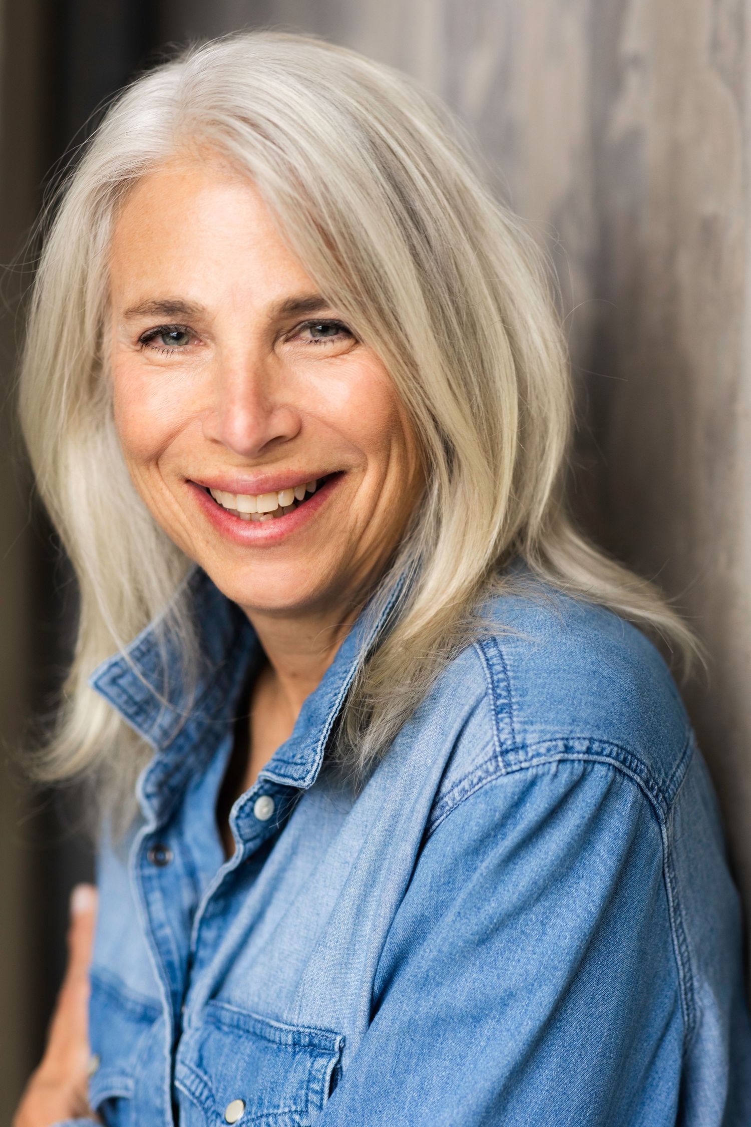 Smiling middle-aged woman with long gray hair wearing a denim shirt, posing against a gray background.