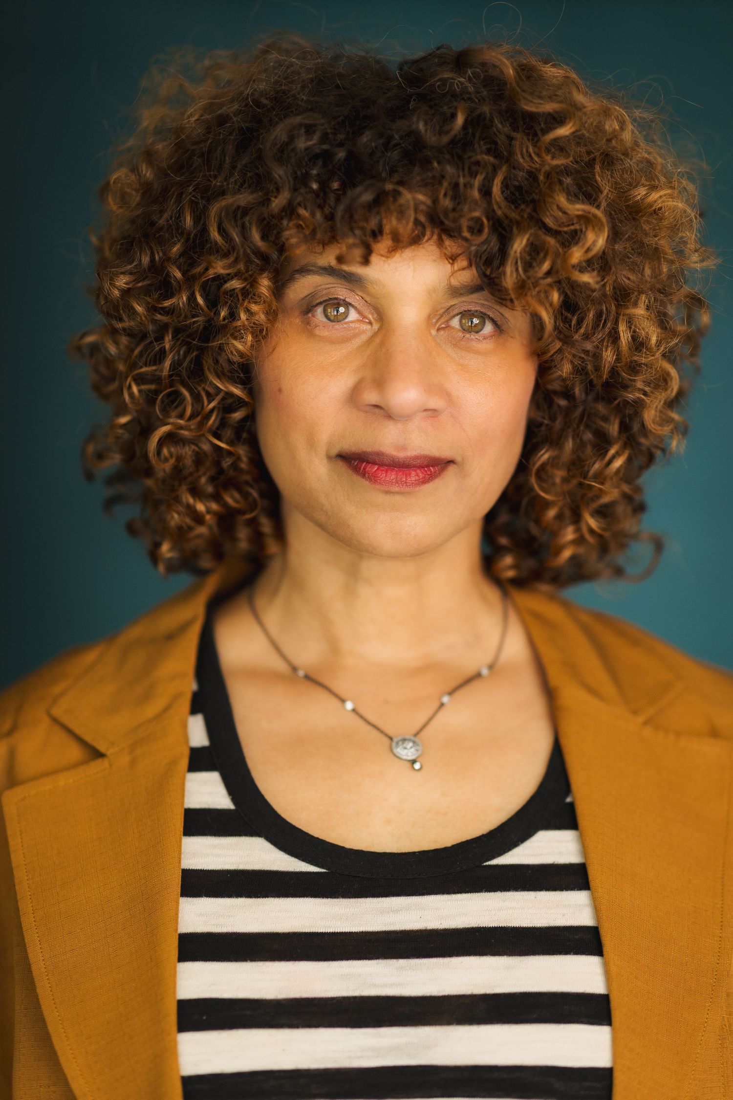 Woman with curly auburn hair wearing a mustard blazer, striped top, and silver necklace against a teal background.