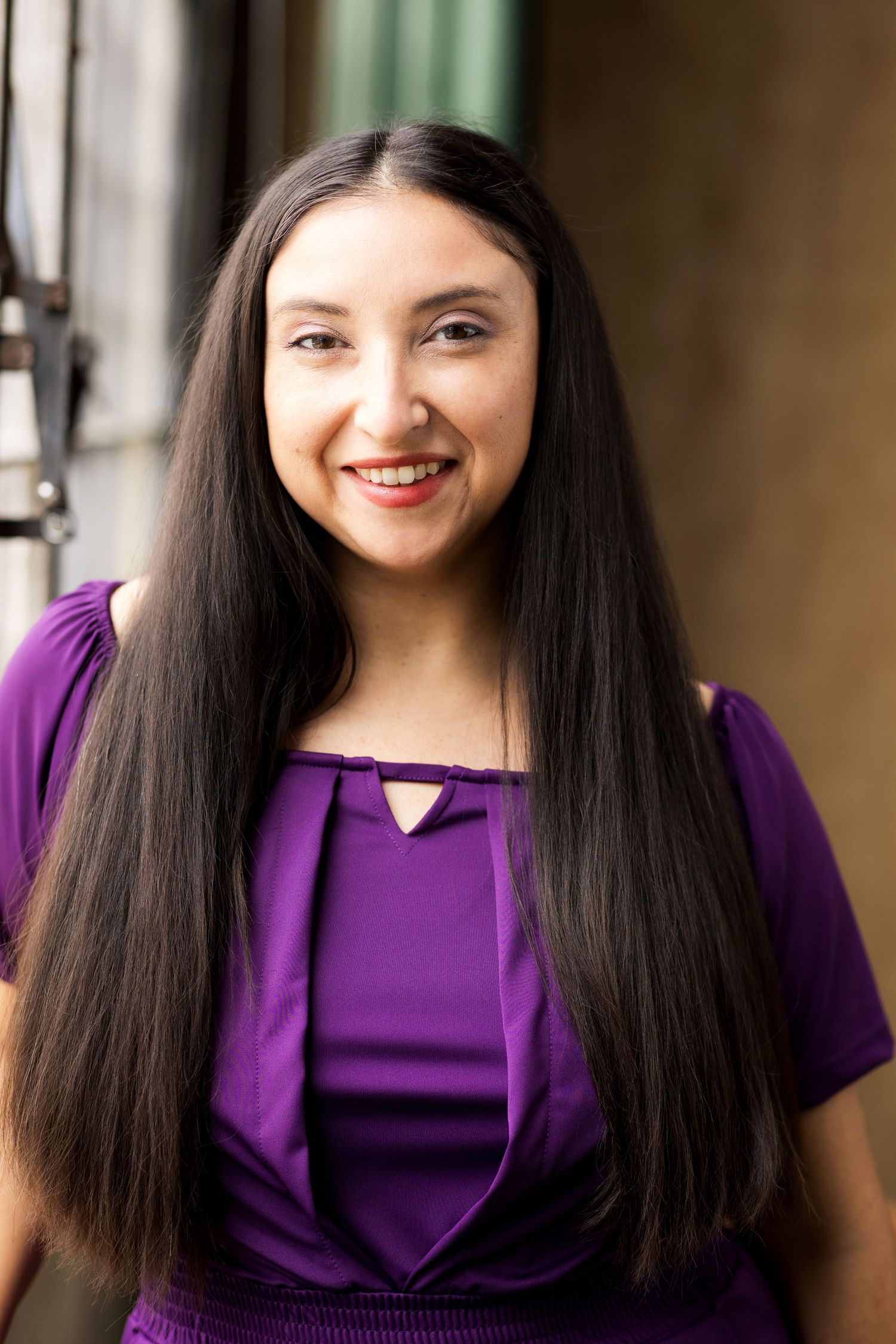 Young woman with long dark hair smiling, wearing a purple top, posed outdoors in a portrait photo.
