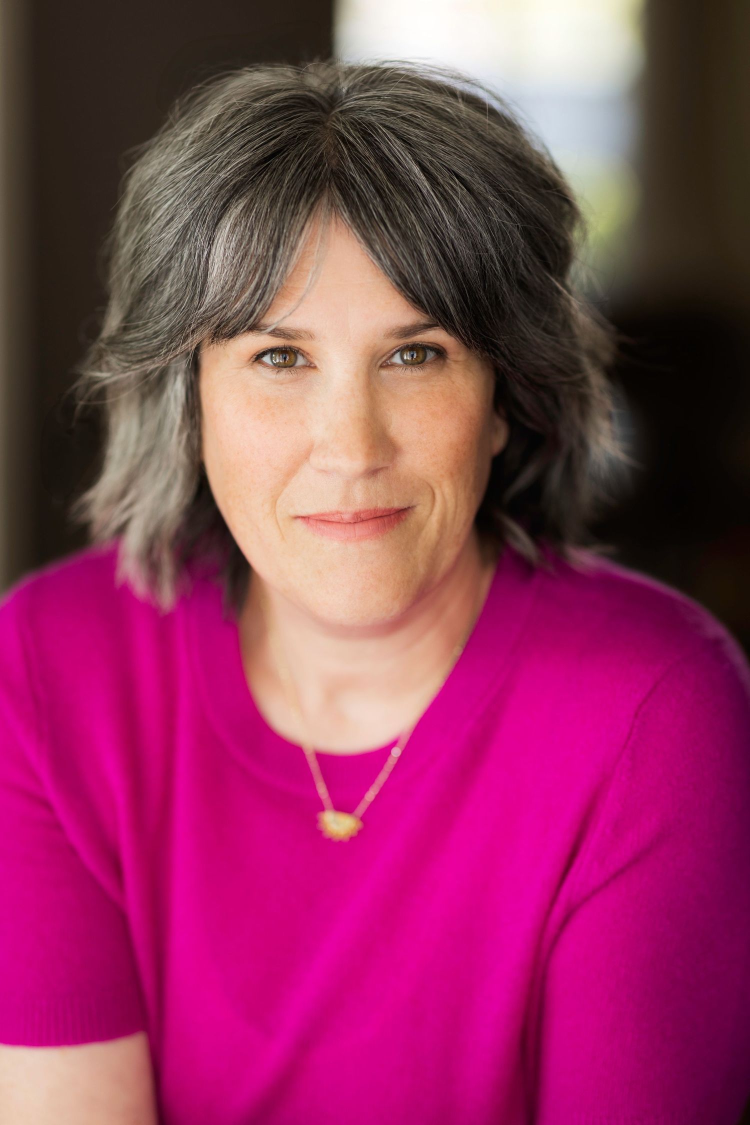 Woman with gray-streaked hair wearing a bright magenta top and gold necklace smiling warmly at the camera.