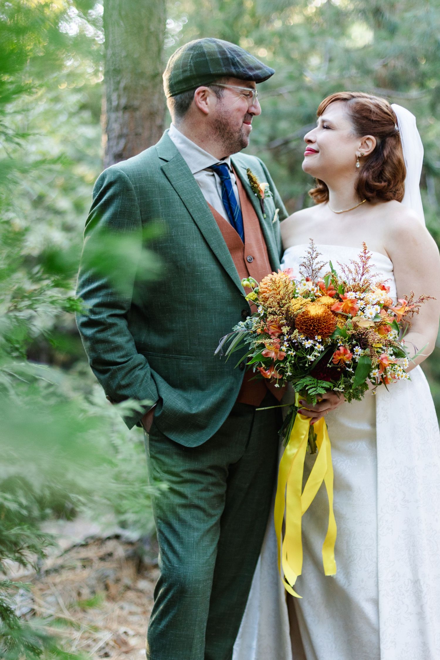 Bride holding autumn-colored bouquet with yellow ribbon next to groom in green suit.