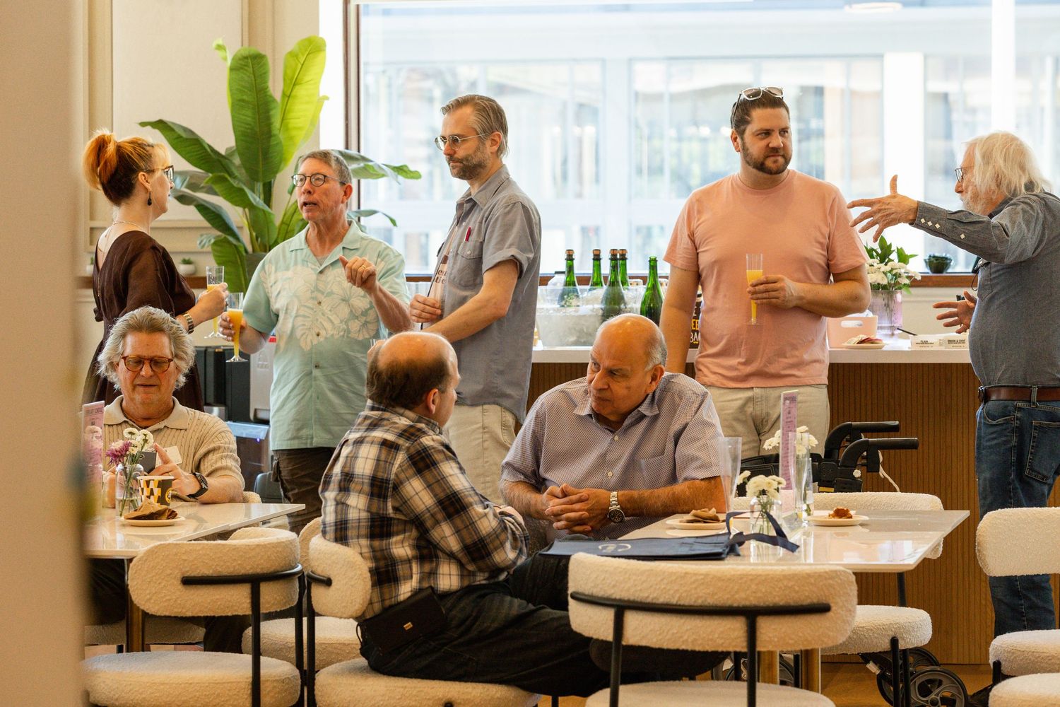 People sitting at round tables in a modern office cafeteria or break room with natural lighting and indoor plants.