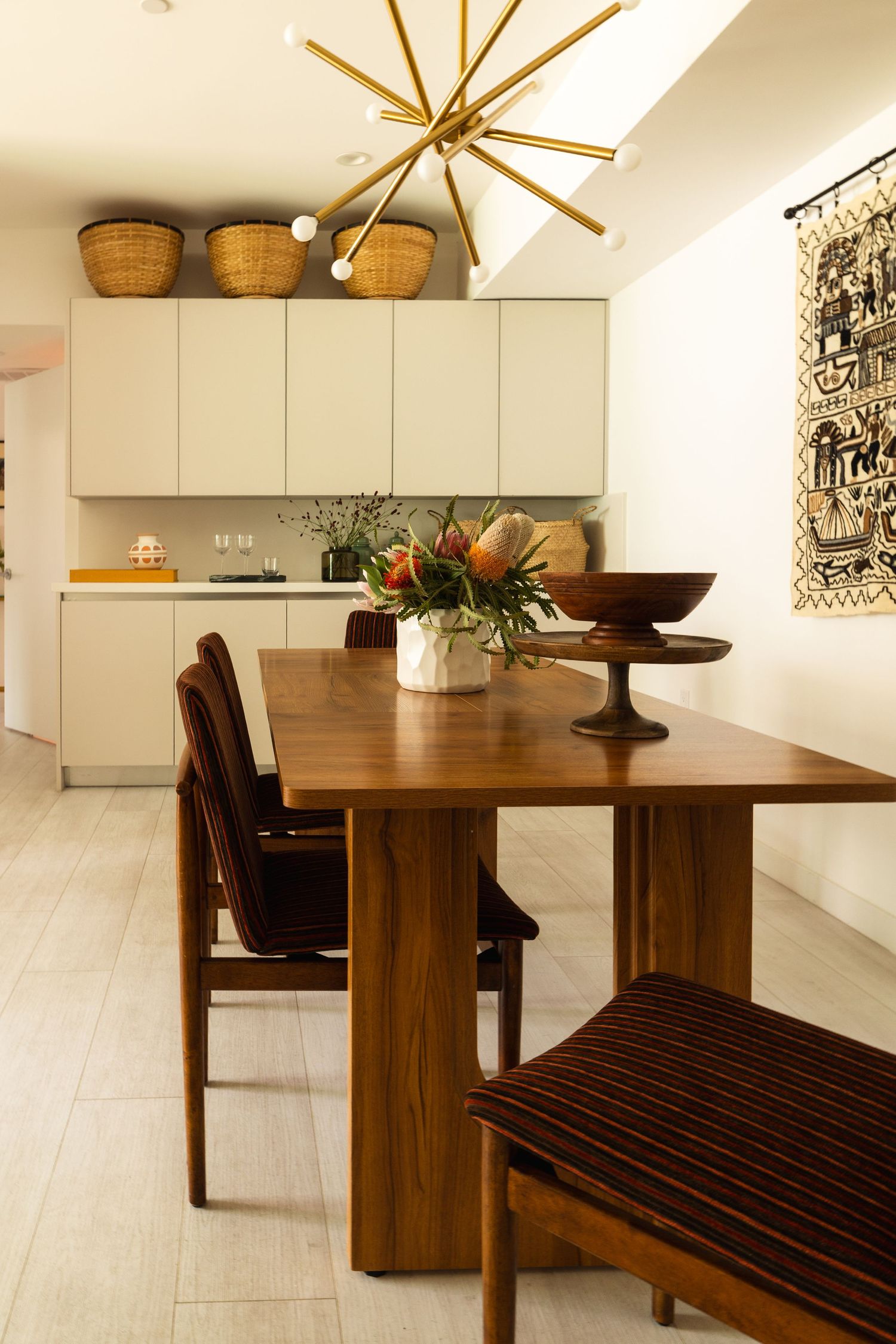 Modern dining area featuring walnut table, brown chairs, gold sputnik light, cream cabinets, and woven baskets above.