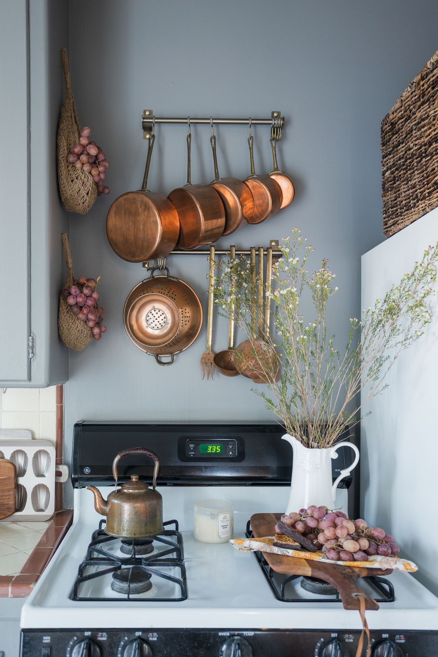 Rustic kitchen with copper pots on brass rack, vintage gas stove, kettle, white pitcher with wildflowers, and grapes on cutting board.