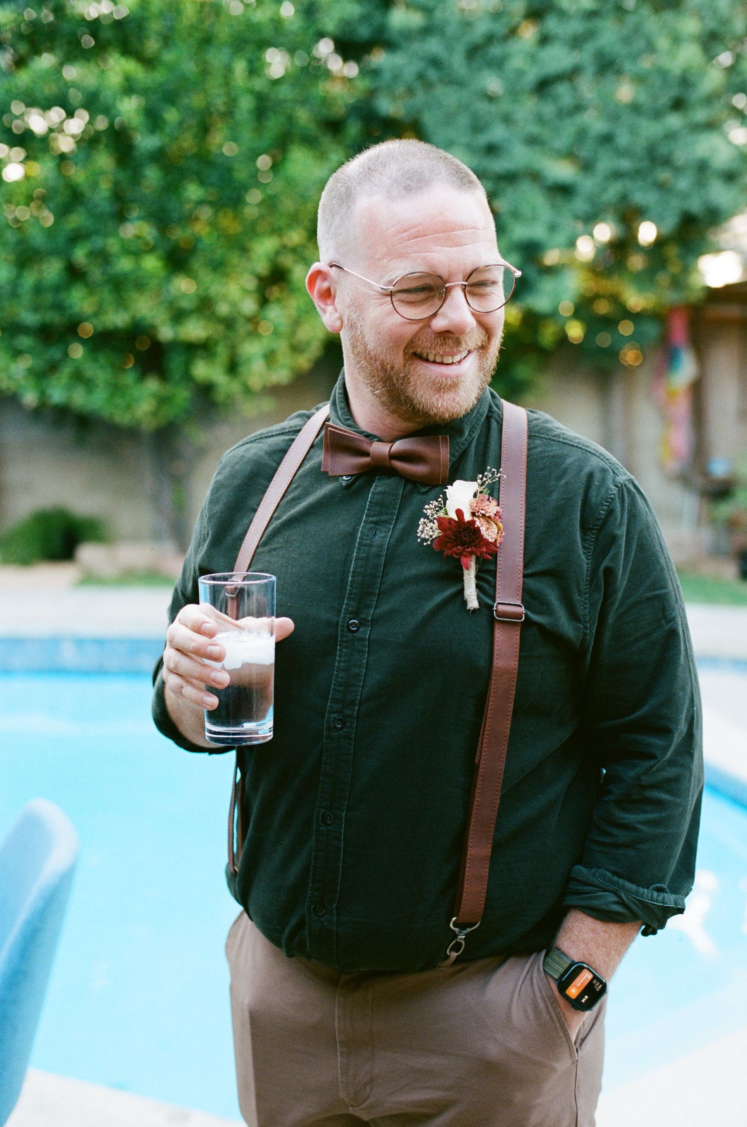 Person in dark outfit with suspenders and bowtie holding drink outdoors.