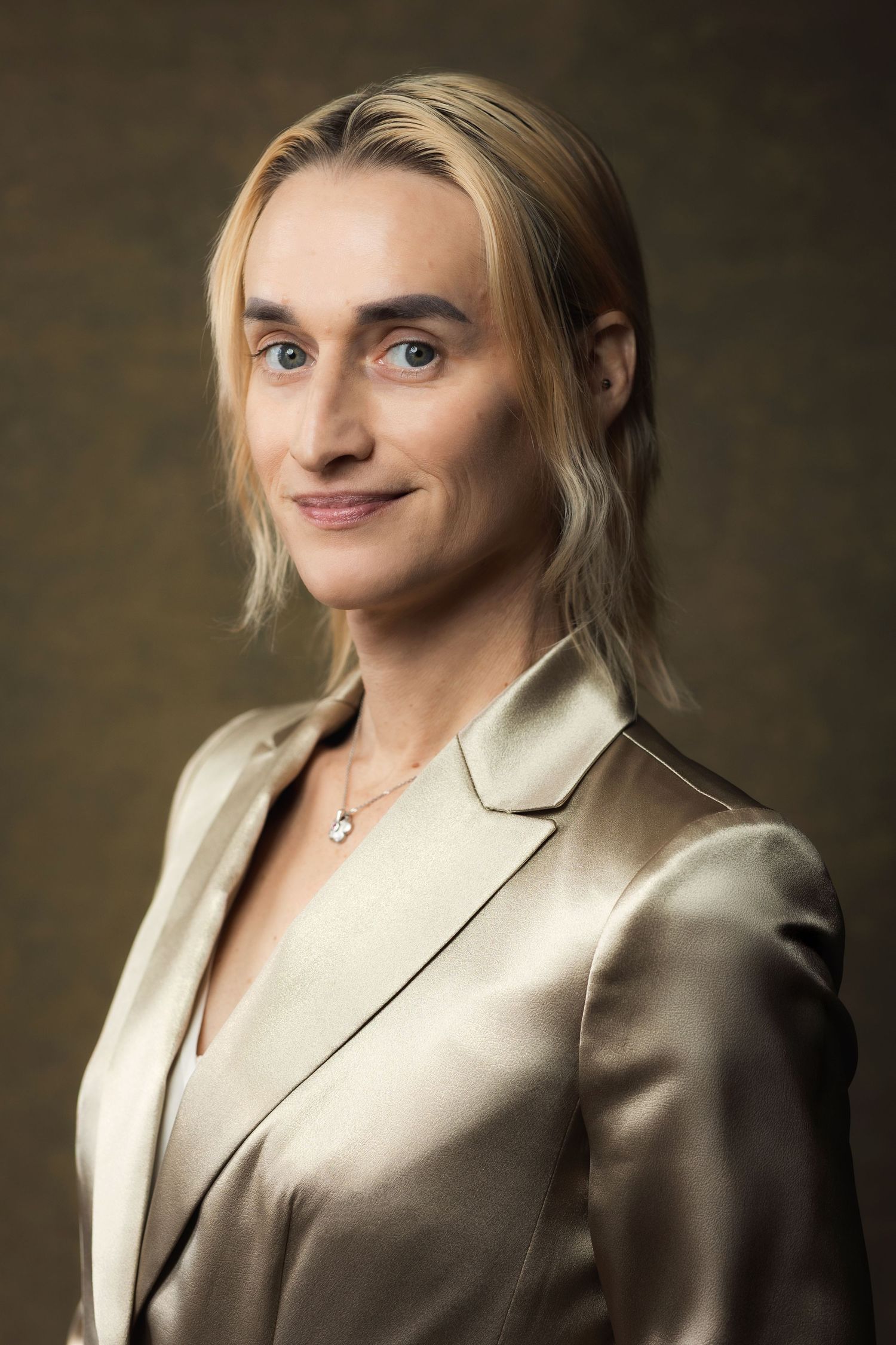 Blonde person in gold satin blazer with necklace, blue eyes, smiling at camera against dark background.