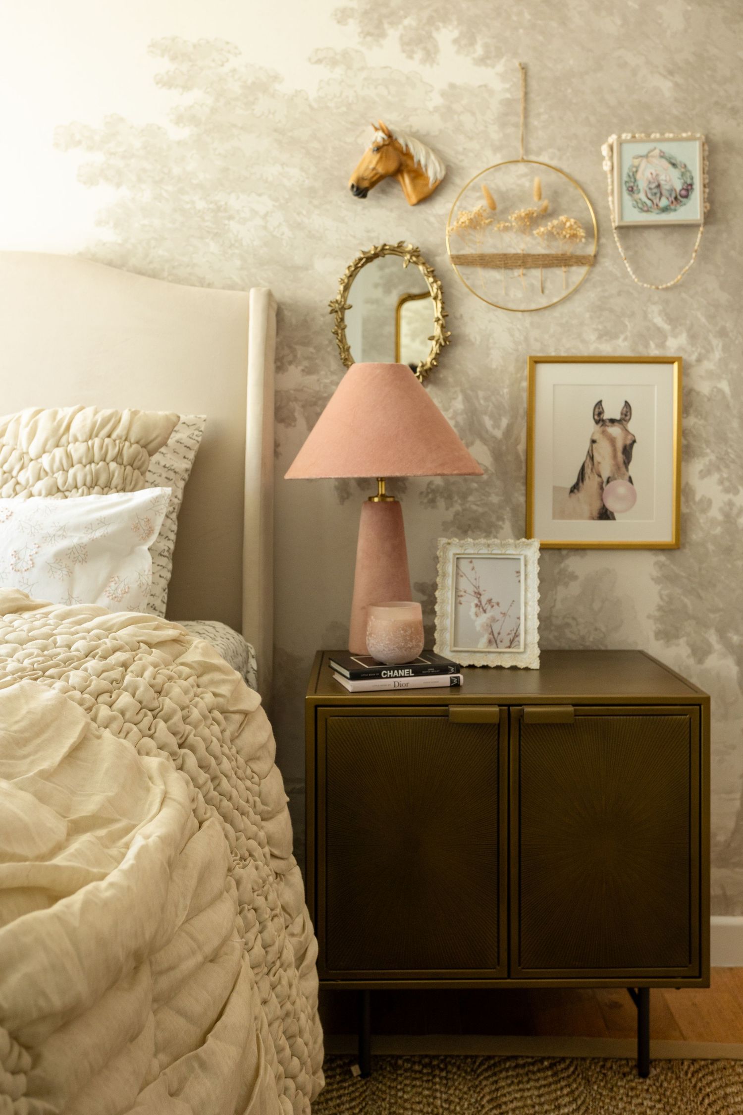 Cozy bedroom with pink lamp, dark nightstand, horse wall art, gold mirror, and textured wallpaper beside a ruffled bedspread.