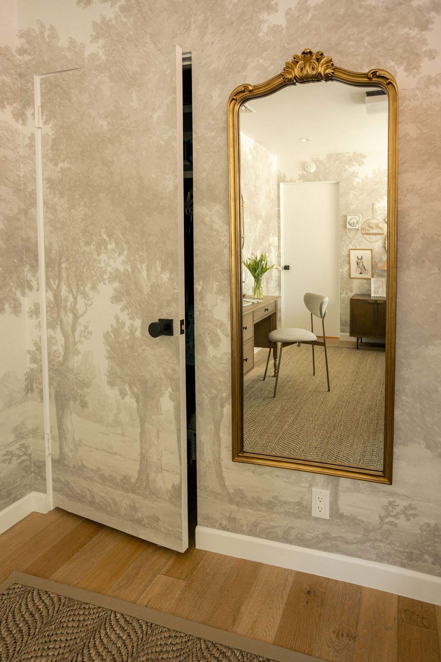 Elegant room with gold-framed floor mirror, toile wallpaper, and hidden door on hardwood floors.