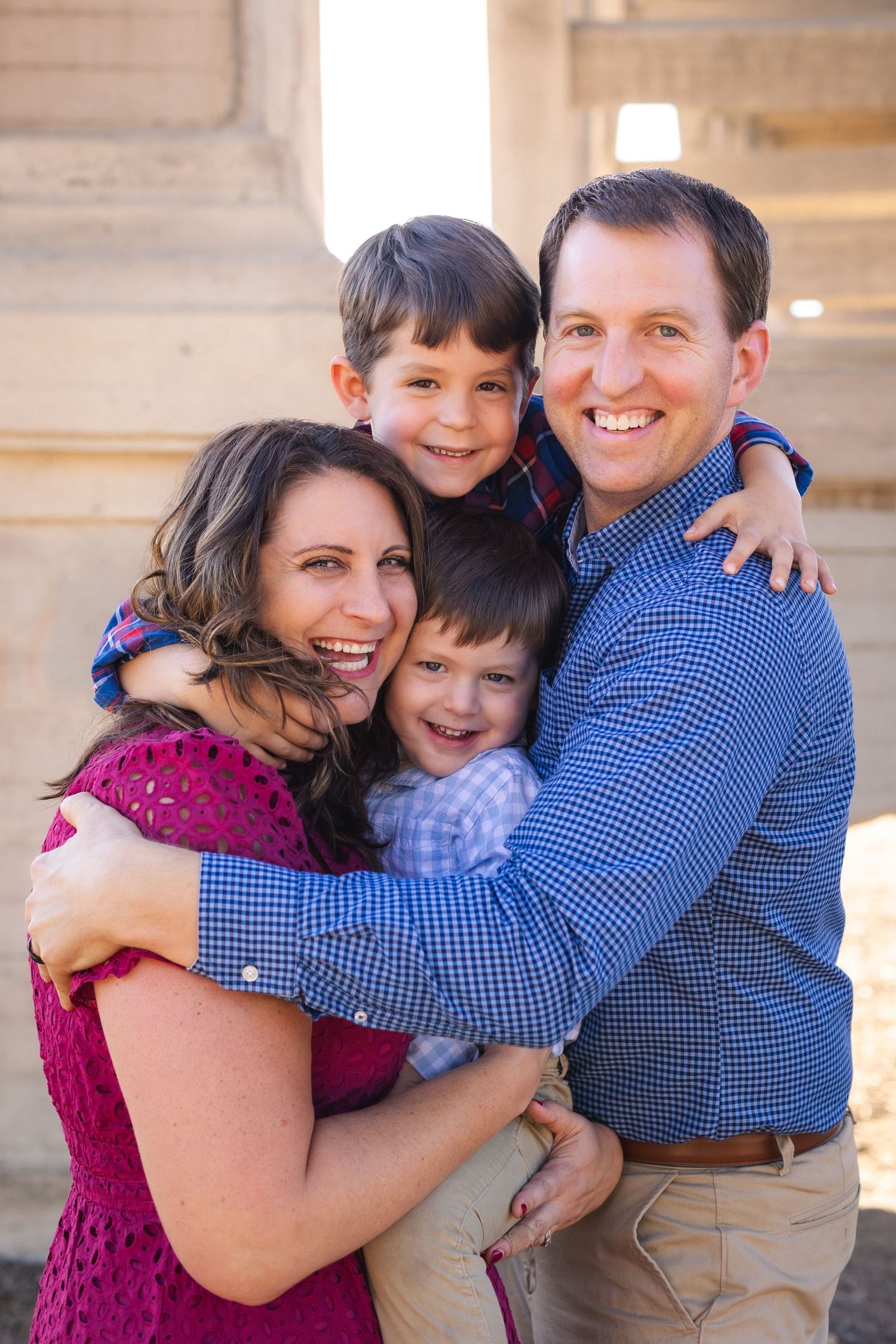Happy family of four hugging and smiling outdoors, parents with two young boys in a warm, sunny setting.