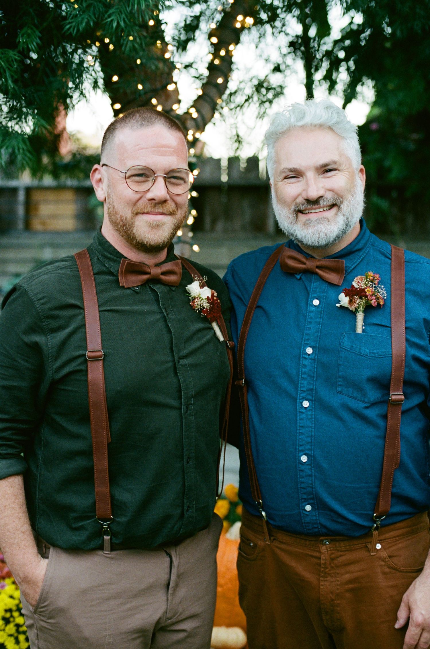 Two people wearing suspenders and button-down shirts standing together in a garden setting.