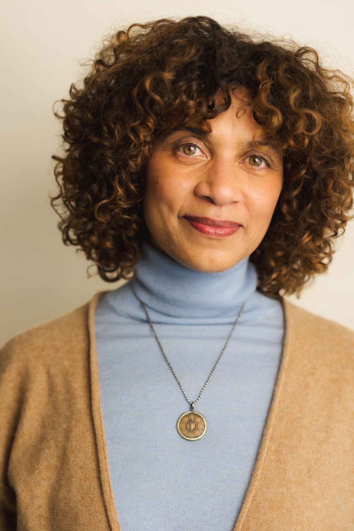 Woman with curly brown hair wearing a blue turtleneck, tan cardigan, and gold medallion necklace smiling softly.