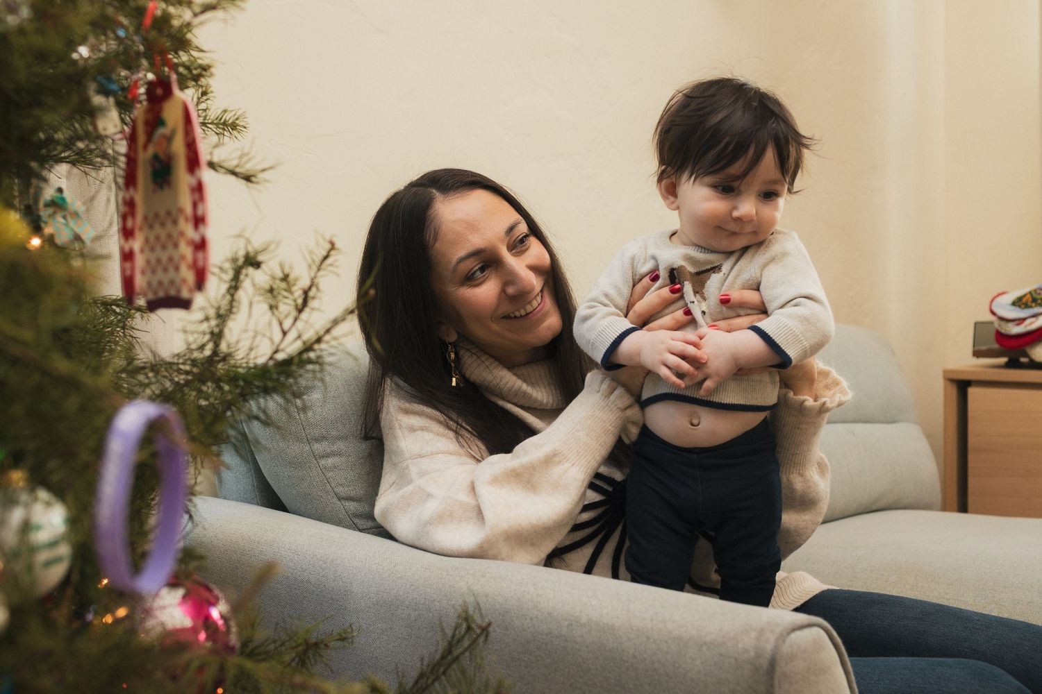 A smiling mother holds her baby boy near a decorated Christmas tree in a cozy living room.