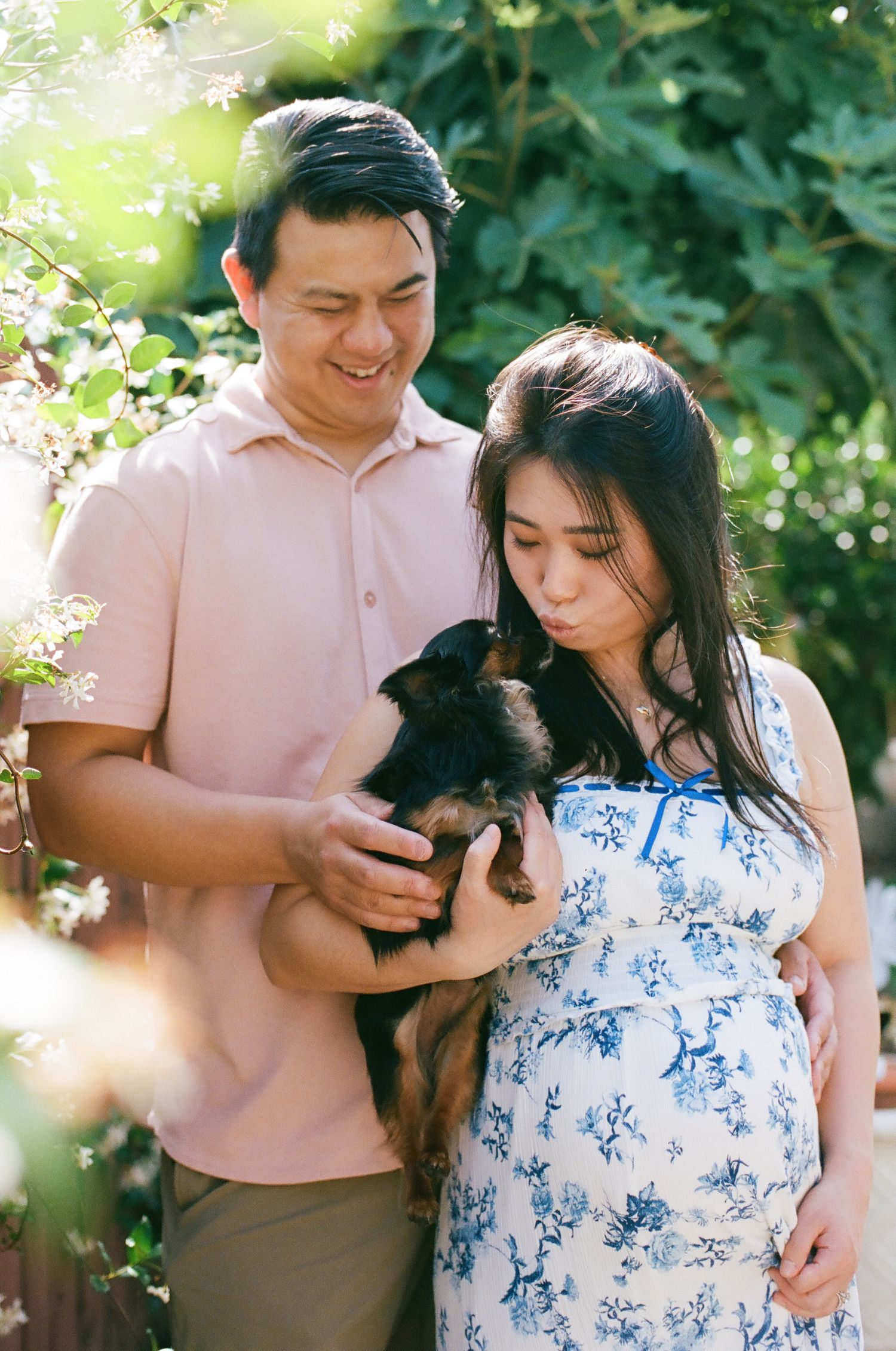 Smiling couple holding a small dog outdoors in a sunny garden, woman in floral dress appears pregnant.