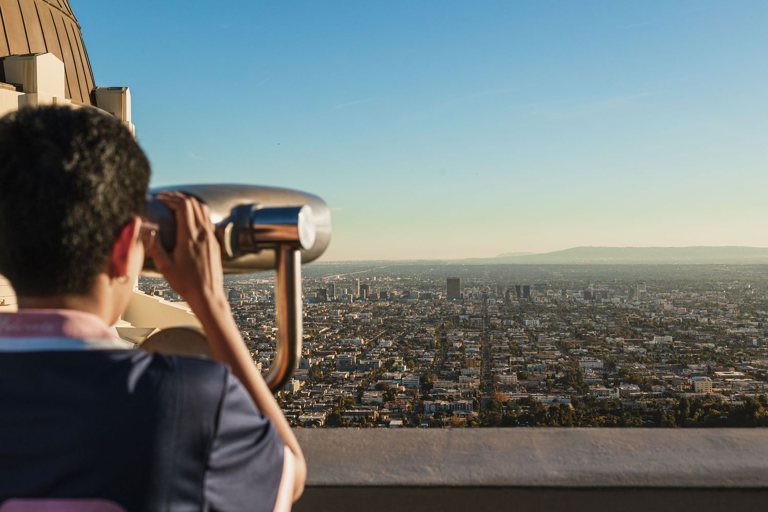 Person using binoculars to view panoramic cityscape from scenic overlook at sunset.