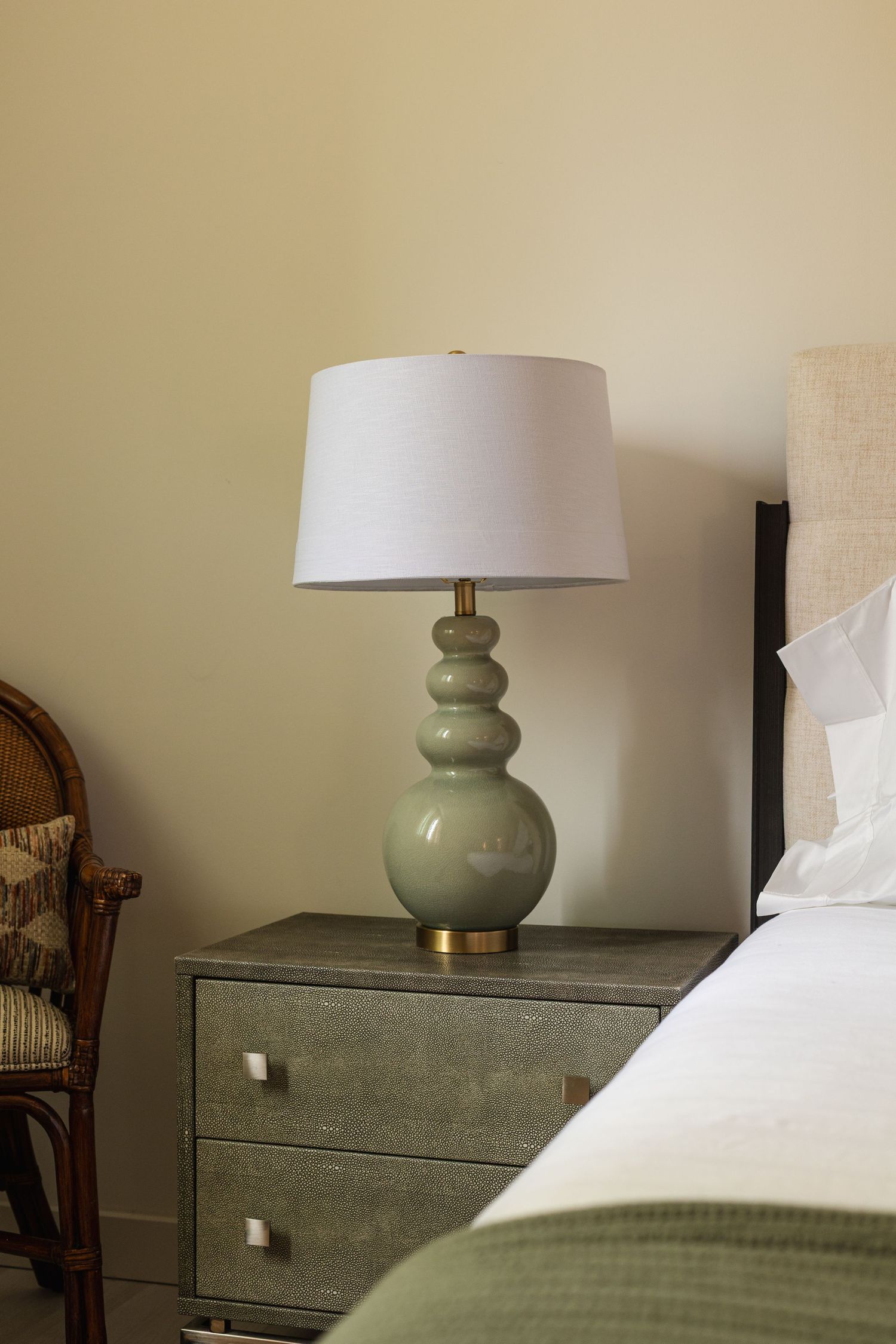 Sage green ceramic table lamp with white drum shade on gray shagreen nightstand beside a bed.