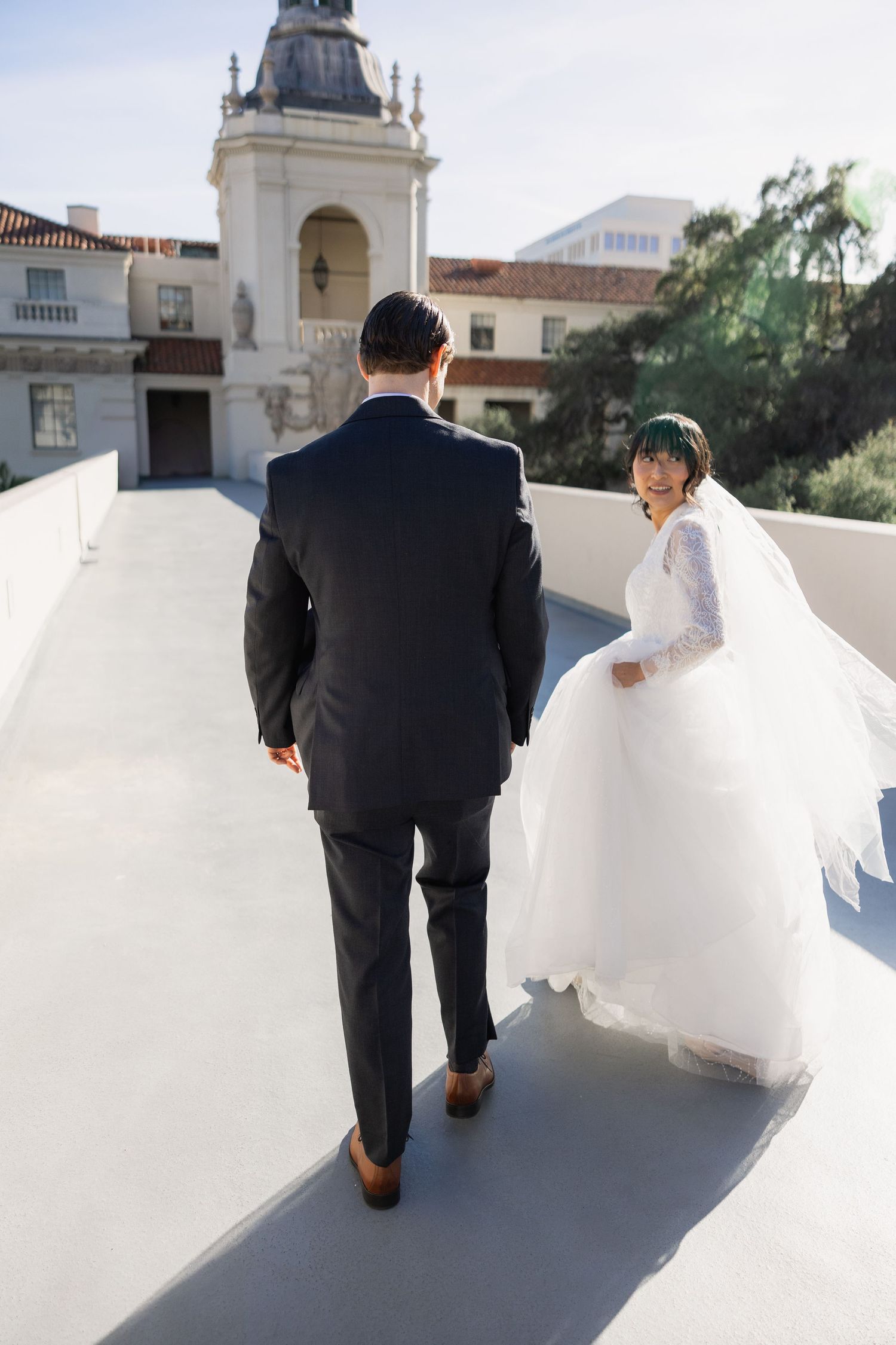 A newlywed couple walking together on a white pathway with Spanish colonial architecture in the background.