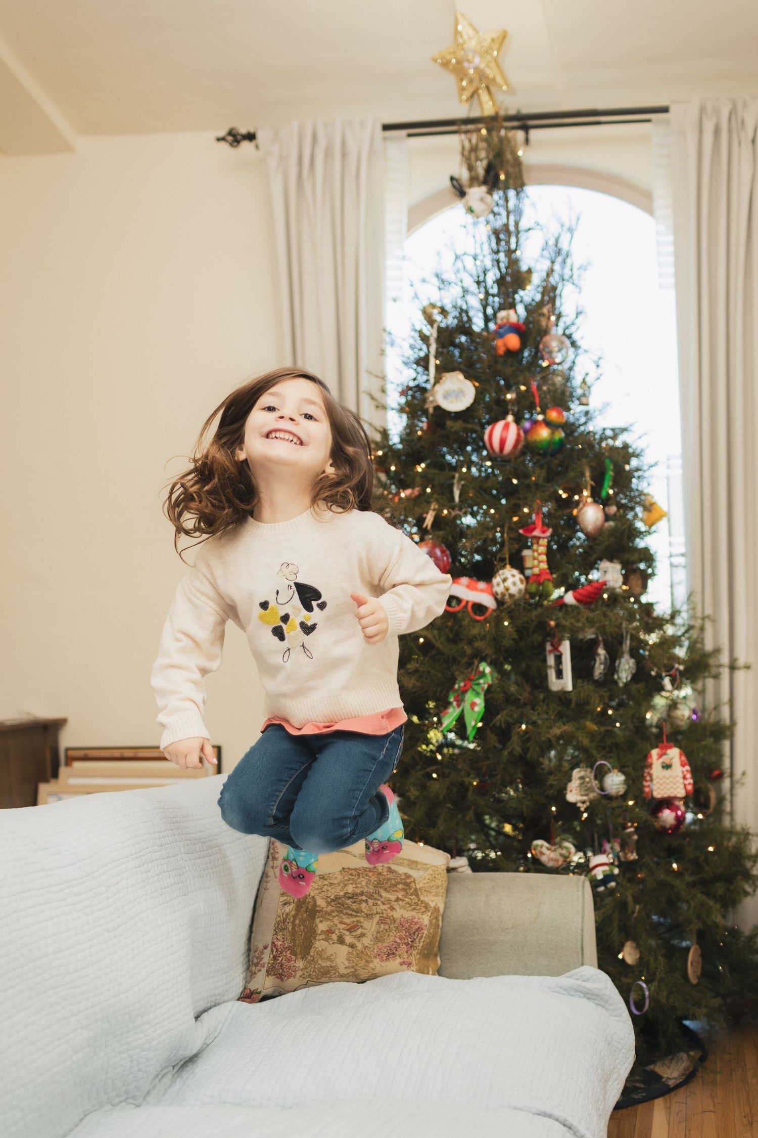 Happy young girl jumping on a sofa in front of a decorated Christmas tree with star topper.