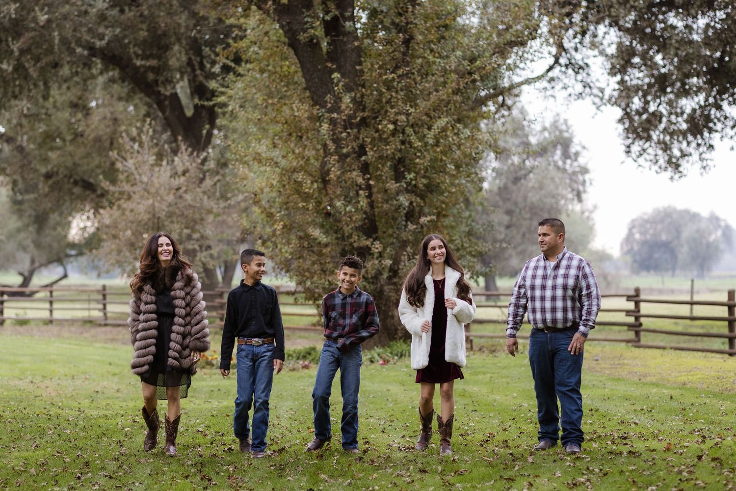 Family of five posing outdoors on green grass near large oak tree and wooden fence on a farm.